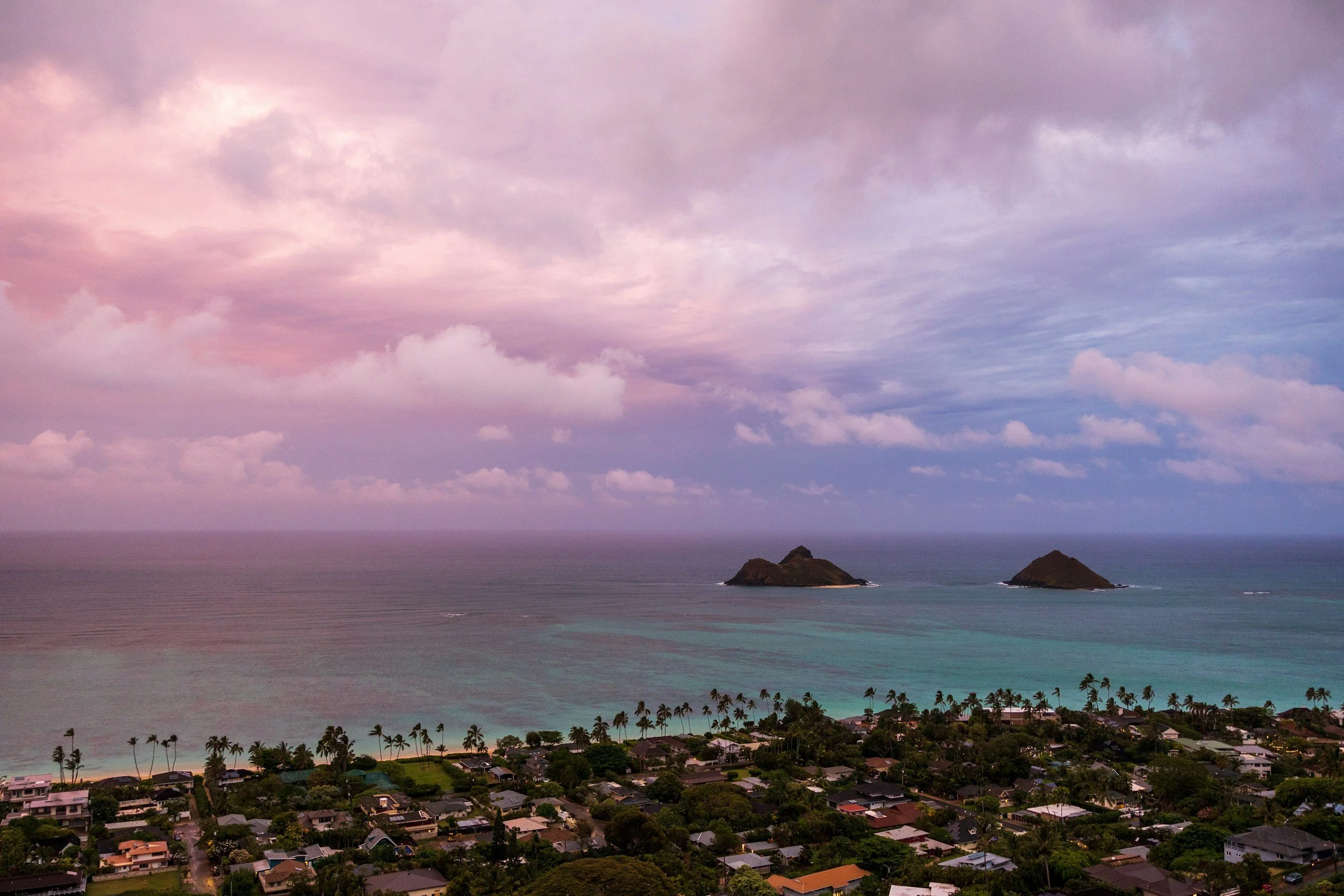 A coastal scene at sunset with a sky filled with pink and purple clouds, two small islands in the ocean, and a residential area with palm trees along the shoreline.