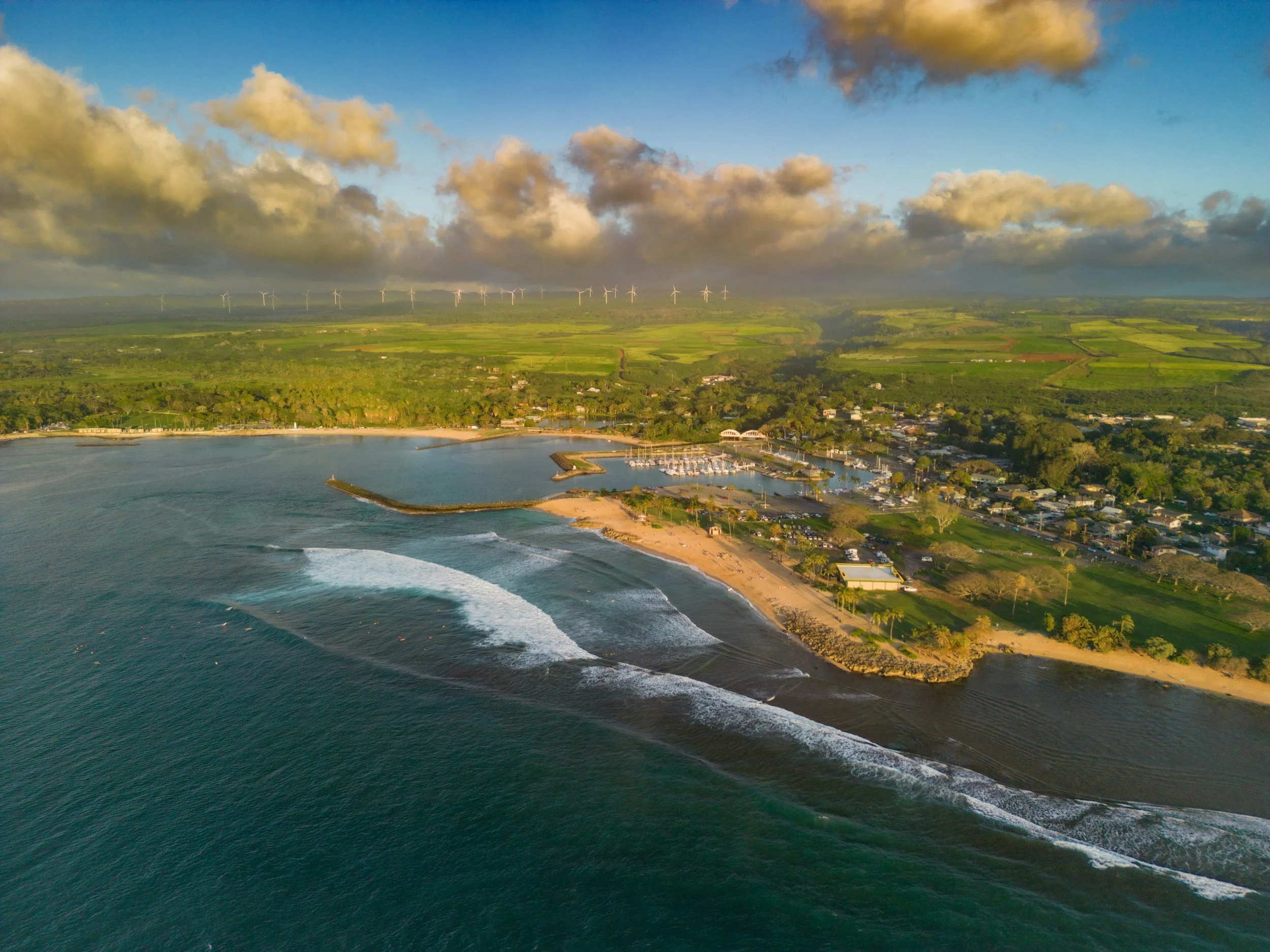 Aerial view of a coastal area during sunset, featuring a sandy beach, a small harbor with boats, residential houses, lush green fields, and wind turbines on the horizon under partly cloudy skies.