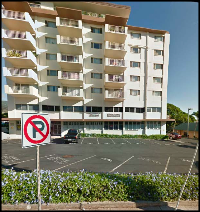 A multi-story residential building with balconies, a parking lot in front, and a no-parking sign in the foreground.