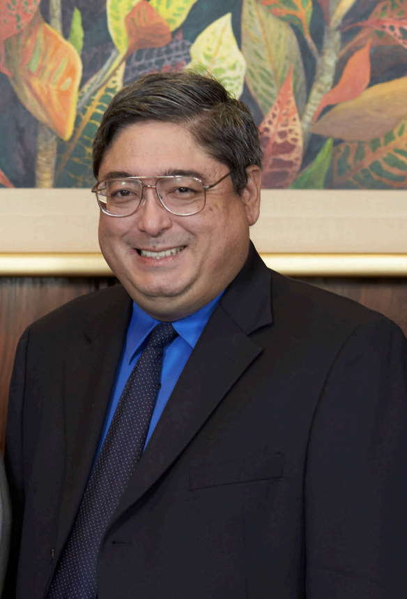 A smiling man with dark hair and glasses wearing a black suit, blue dress shirt, and dotted tie, standing in front of a colorful plant-themed painting.