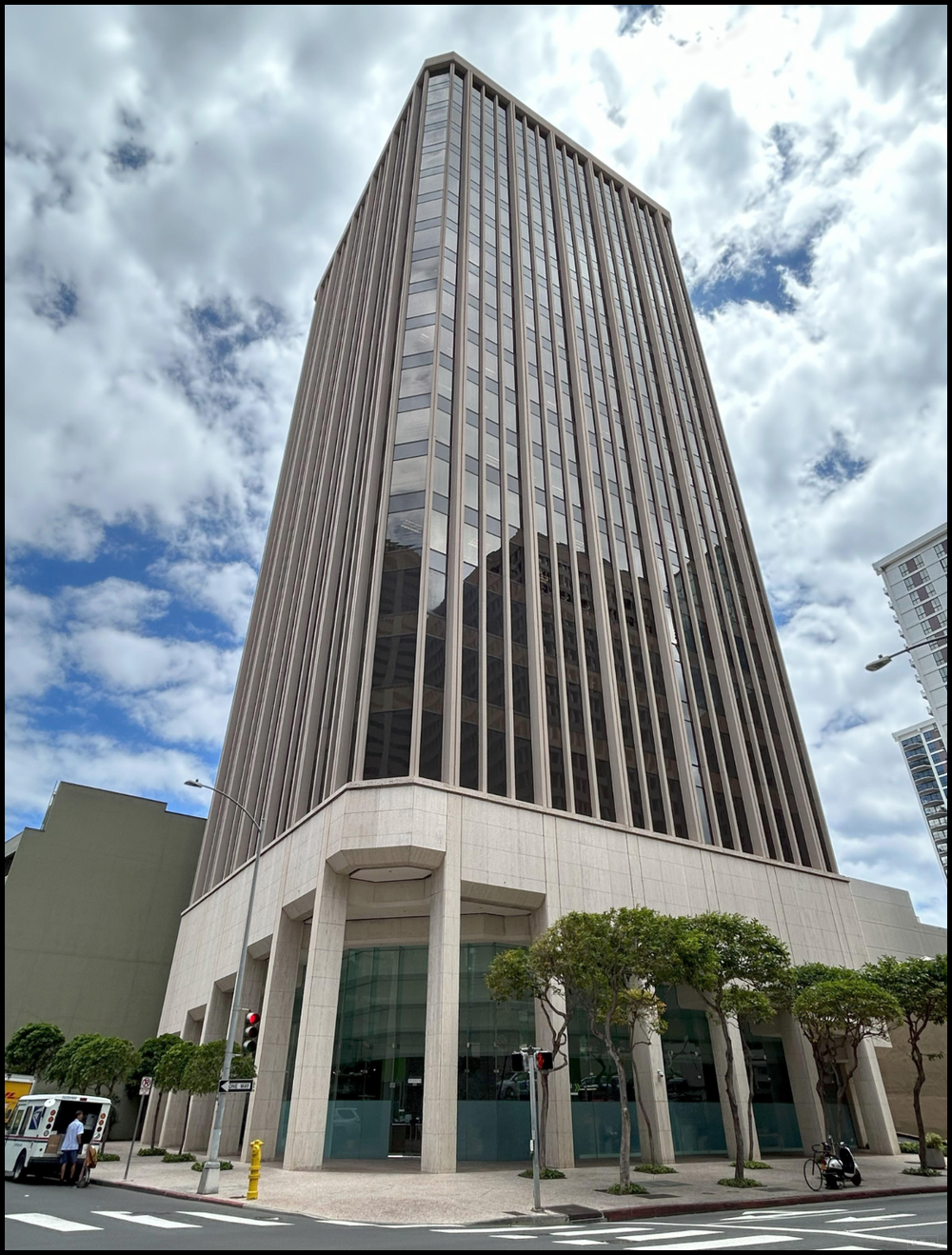 Tall modern office building with glass windows and vertical metal panels, surrounded by trees and city streets under partly cloudy sky.