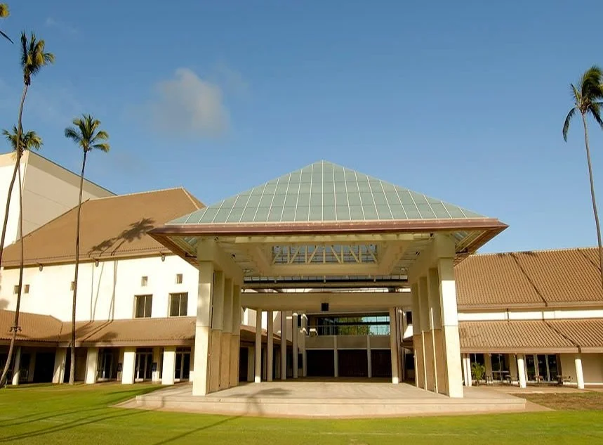 A modern building with a glass pyramid roof, surrounded by palm trees and a well-maintained lawn, under a clear blue sky.