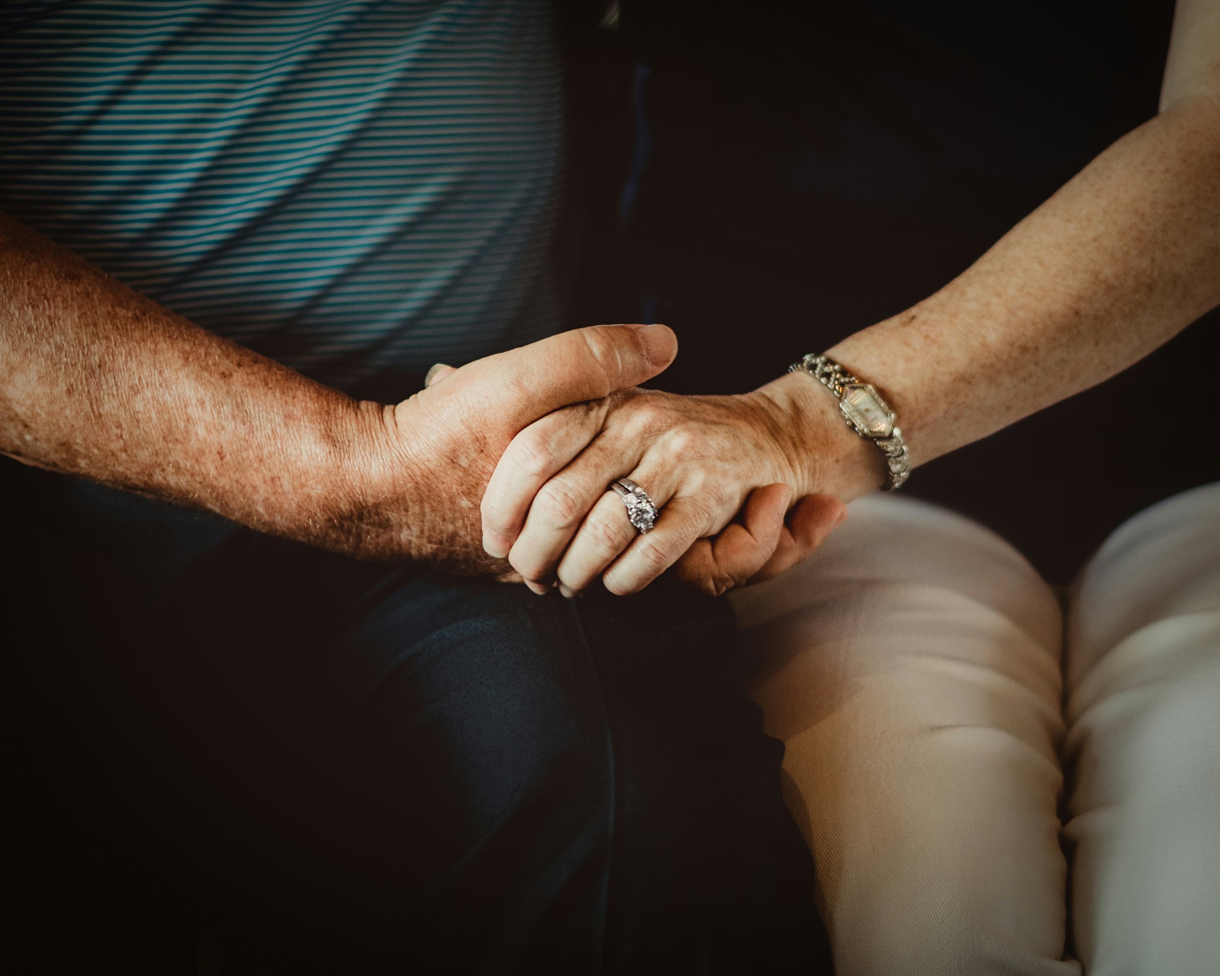 Close-up of an elderly couple holding hands, showing their wedding rings and jewelry, with one person's arm affecting a lot of freckles.