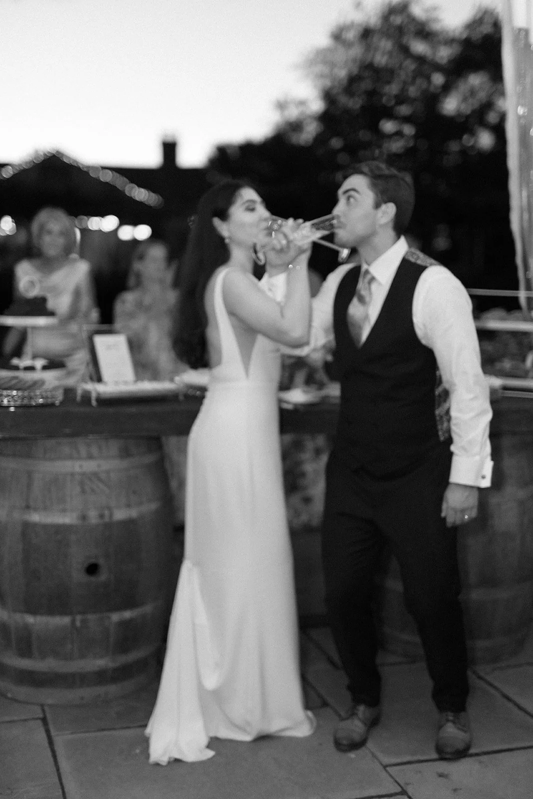 Black and white photo of a bride and groom drinking from a champagne bottle at their wedding reception, with a bar and guests in the background.