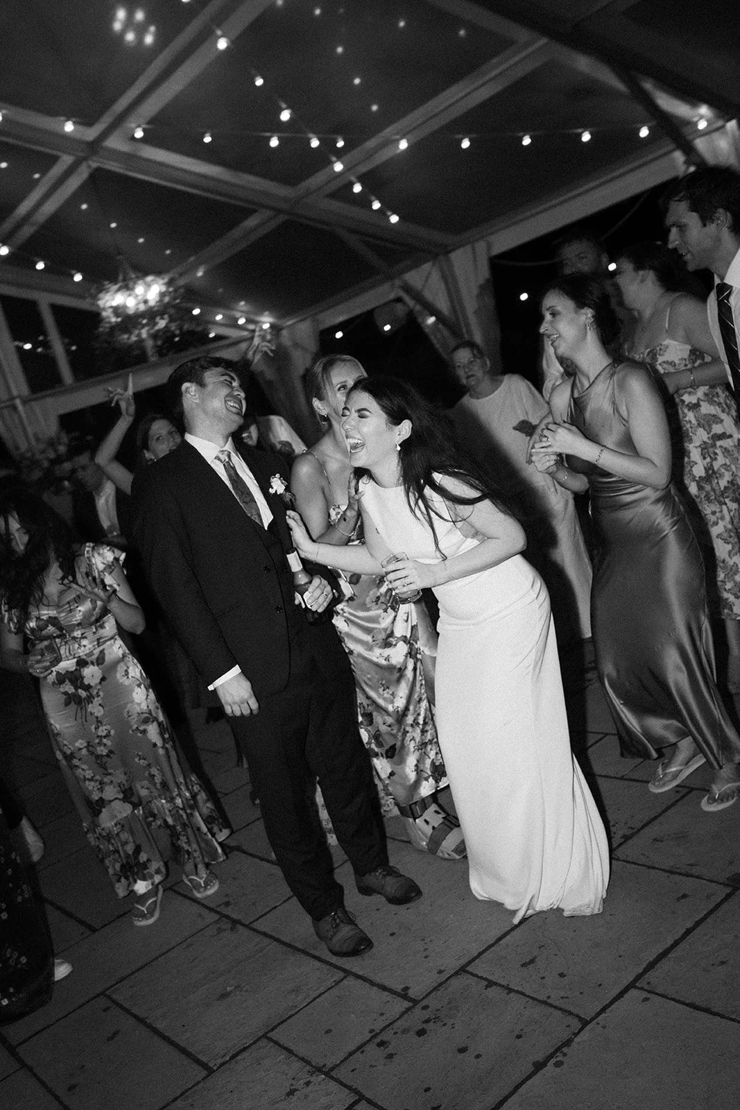 People dancing and celebrating at a wedding reception under string lights, with the bride and groom in the center laughing and enjoying the party.