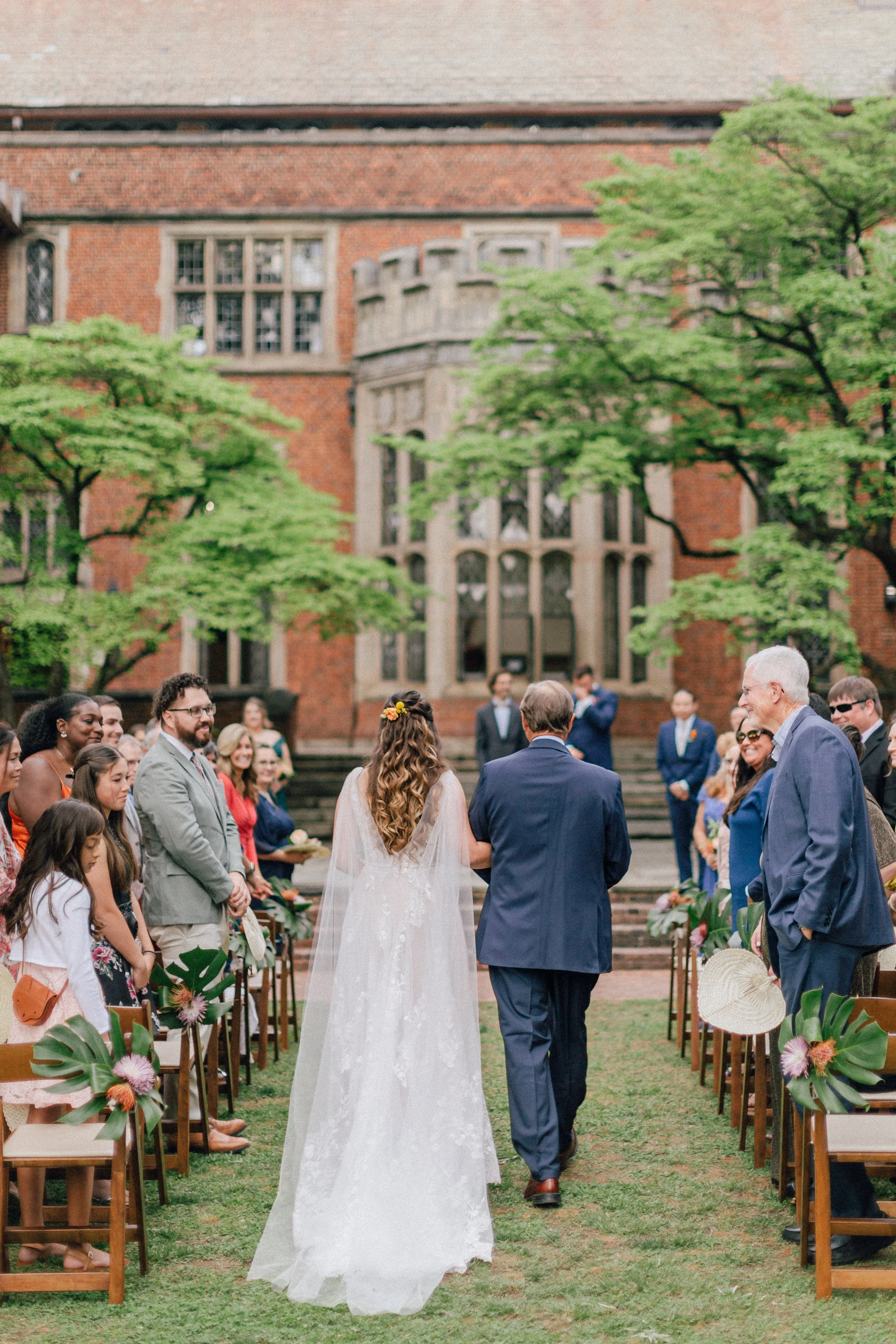 A bride walking down the aisle at an outdoor wedding ceremony, surrounded by guests seated on either side, with a historic brick building and green trees in the background.