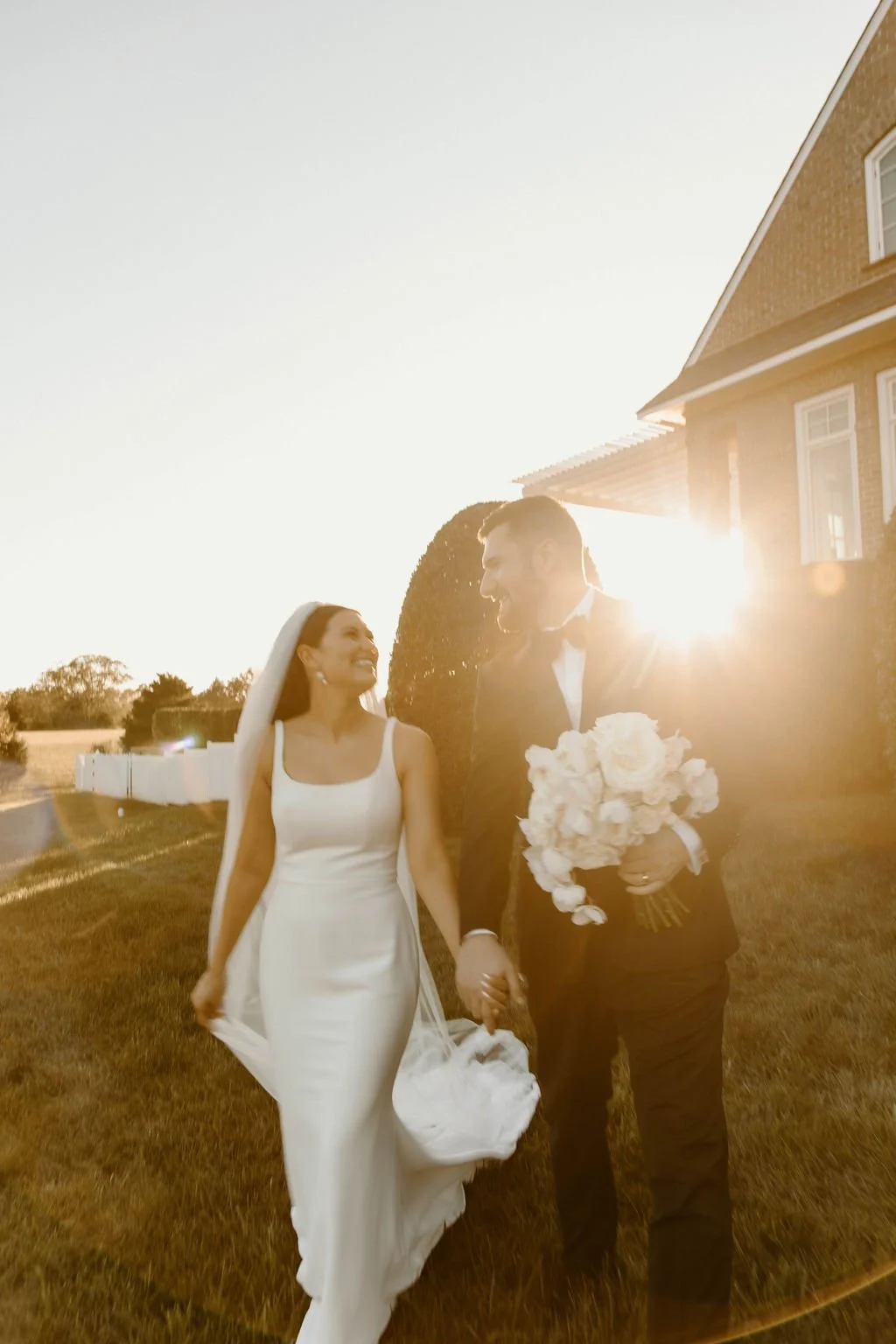 Bride and groom walking hand in hand outside during sunset, with the bride in a white wedding dress and veil, and the groom in a black tuxedo holding a bouquet of white roses.