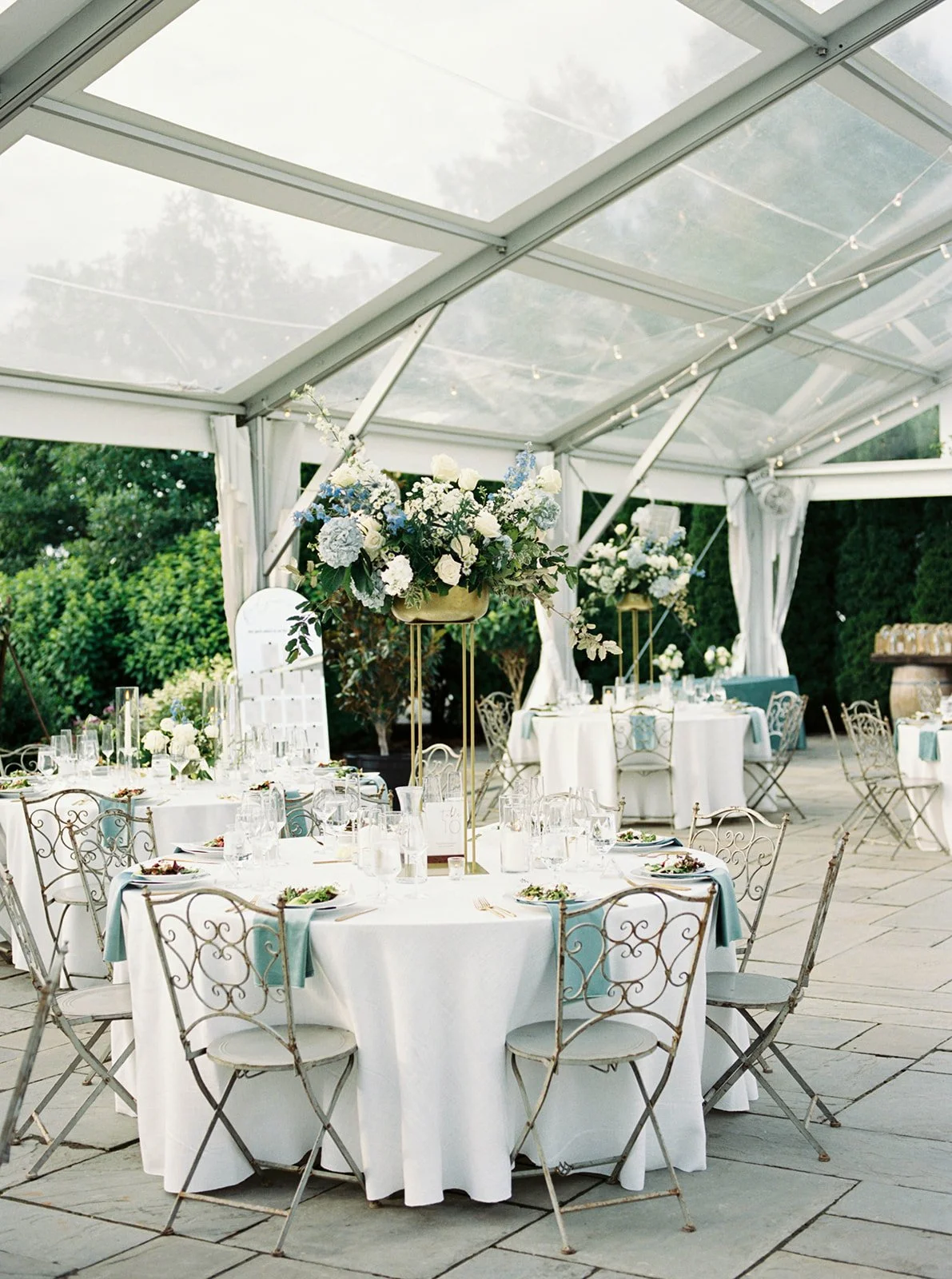 Elegant outdoor wedding reception under a white tent with round tables decorated with white tablecloths, floral centerpieces featuring white and blue flowers, and gold accents, set for dining with plates, glasses, and cutlery.