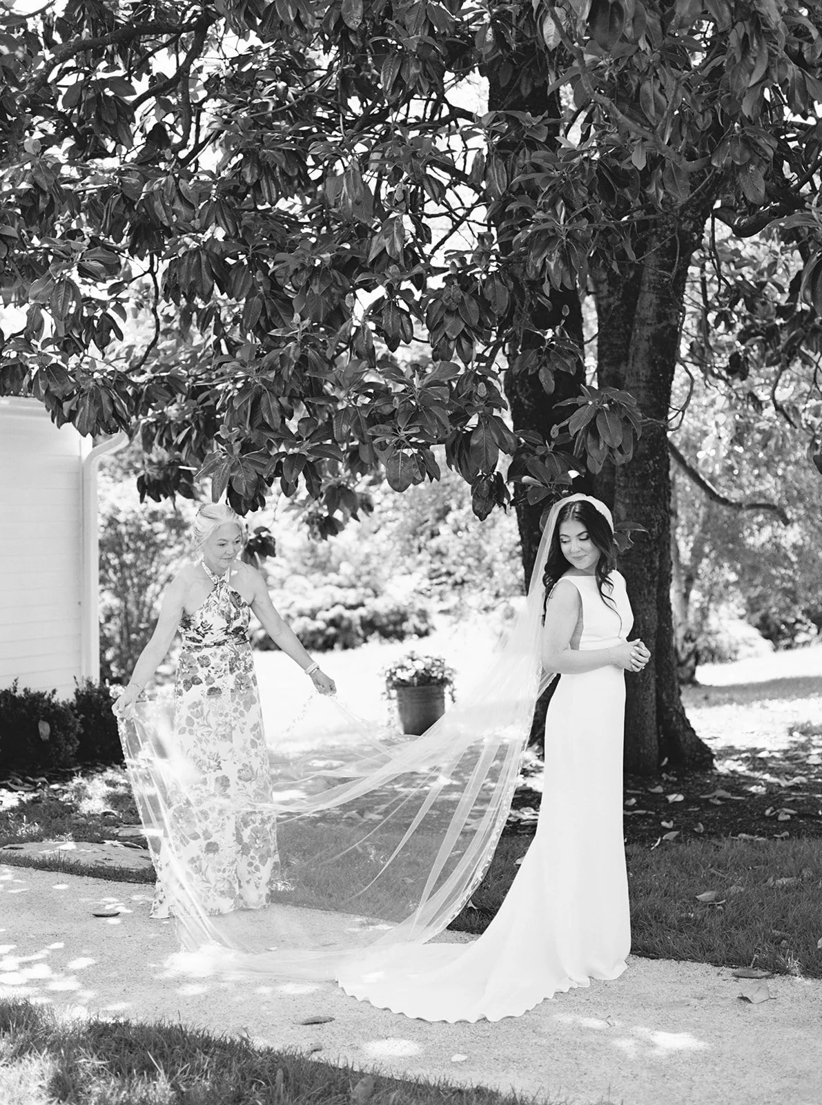 A bride in a white wedding dress and veil outdoors, with an older woman assisting her with her veil under a large tree, in a garden setting.