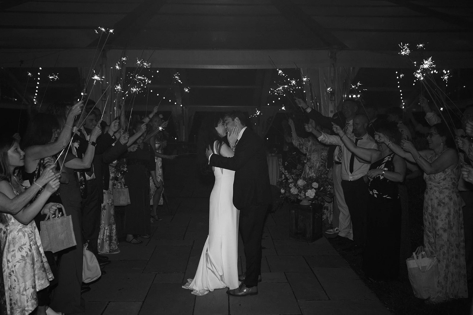 A wedding couple sharing a kiss under sparklers held by friends and family in a celebration at night.