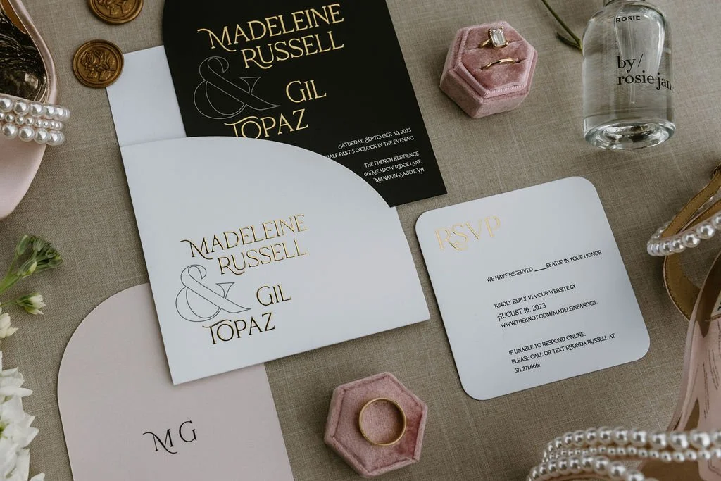 Wedding invitation suite with black and white cards, RSVP card, pink velvet ring box with a ring, glass perfume bottle labeled Rosie by Rosie Jane, and gold rings on a table.