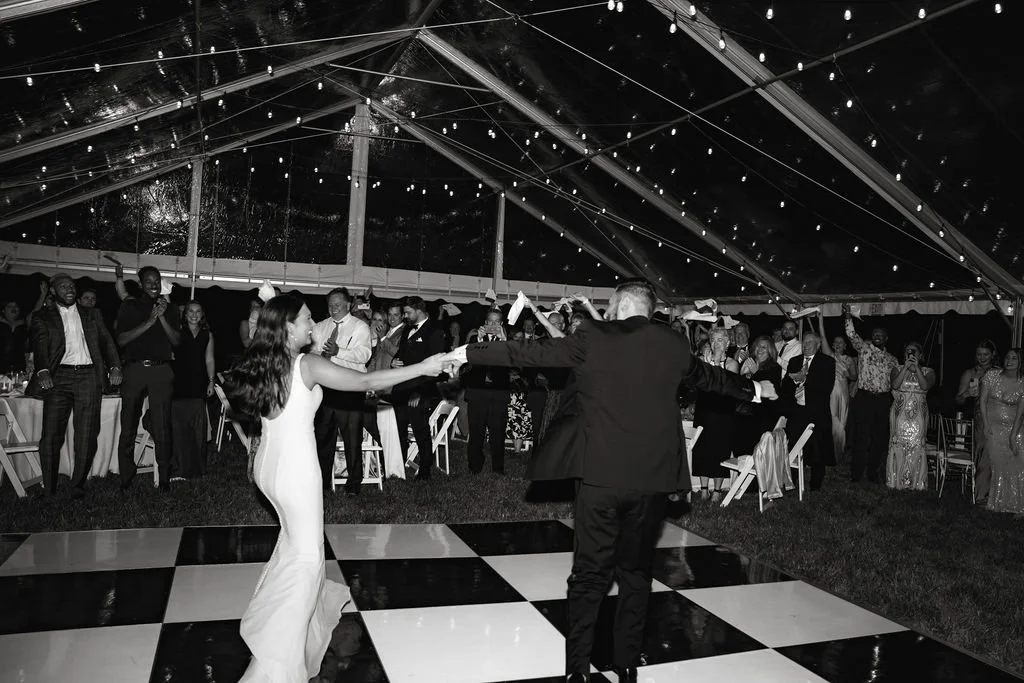 Couple dancing in wedding reception under string lights, guests watching in the background, black and white photograph.