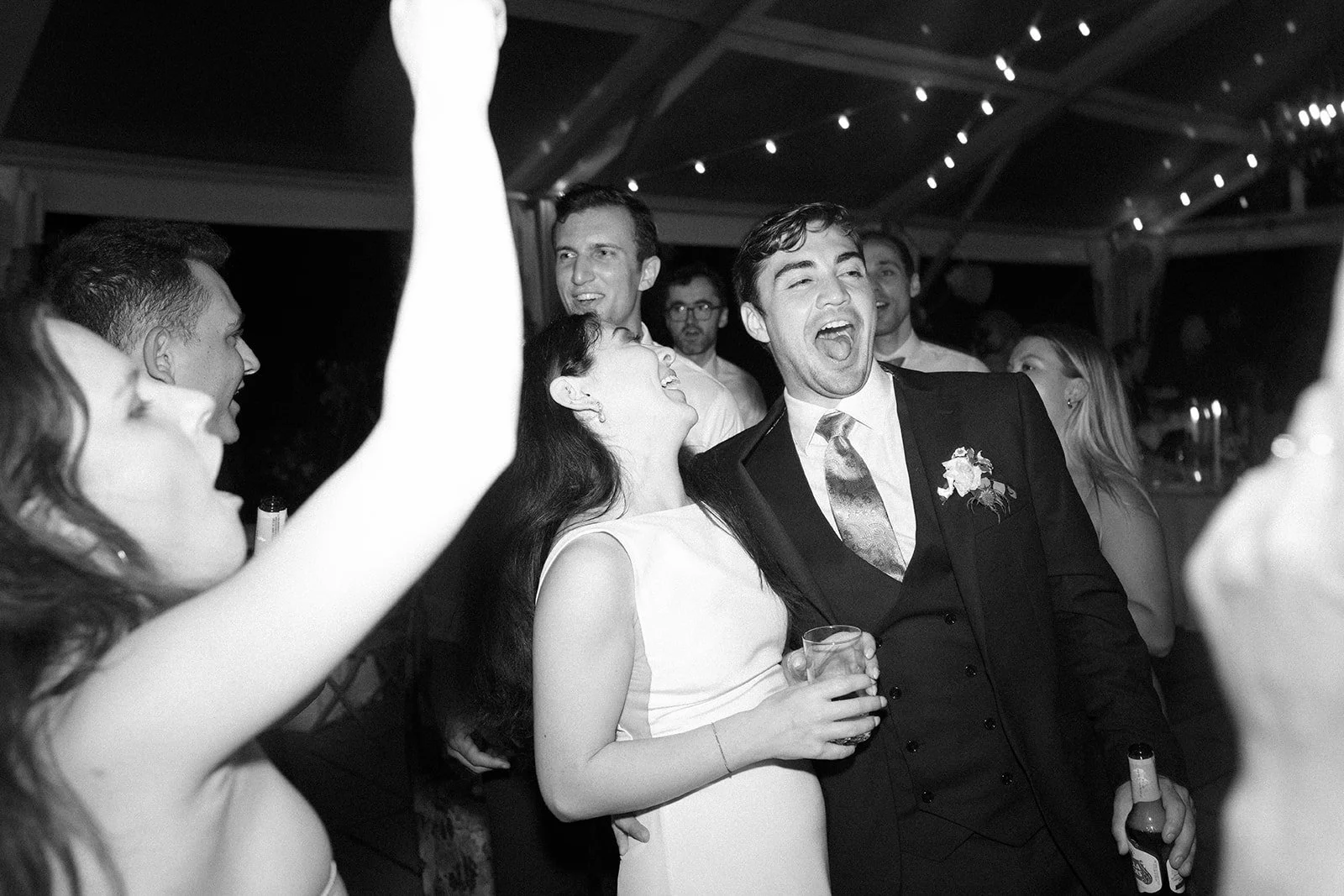 People partying at a celebration, smiling and laughing, some holding drinks, in a decorated venue with string lights on the ceiling.