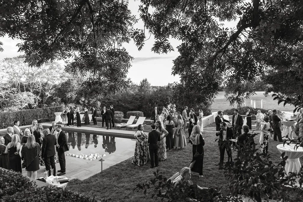 People socializing and mingling at a formal outdoor event by a pool, with trees and open fields in the background.