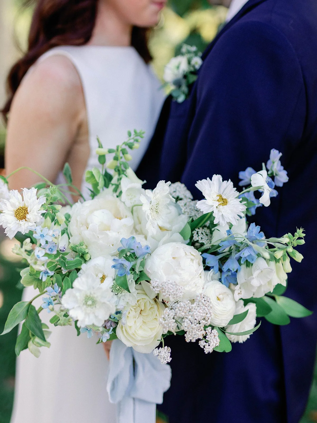 Close-up of a large white and blue flower bouquet held by a person in a formal suit, with a woman in a white dress standing behind them, at an outdoor event.