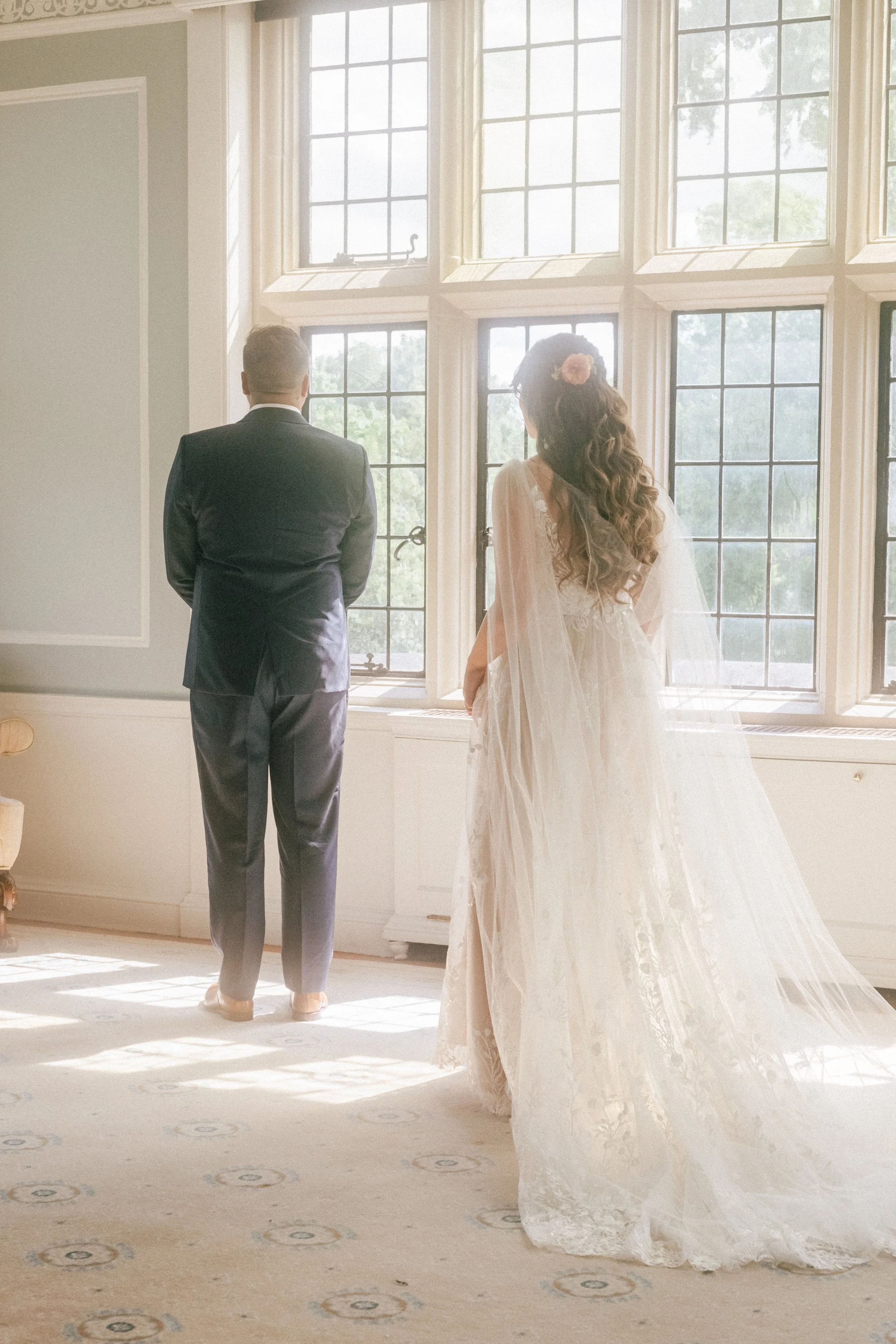 Bride and groom standing near a large window, facing away from the camera, during a wedding ceremony. The bride wears a lace wedding dress with a veil, and the groom wears a dark suit.