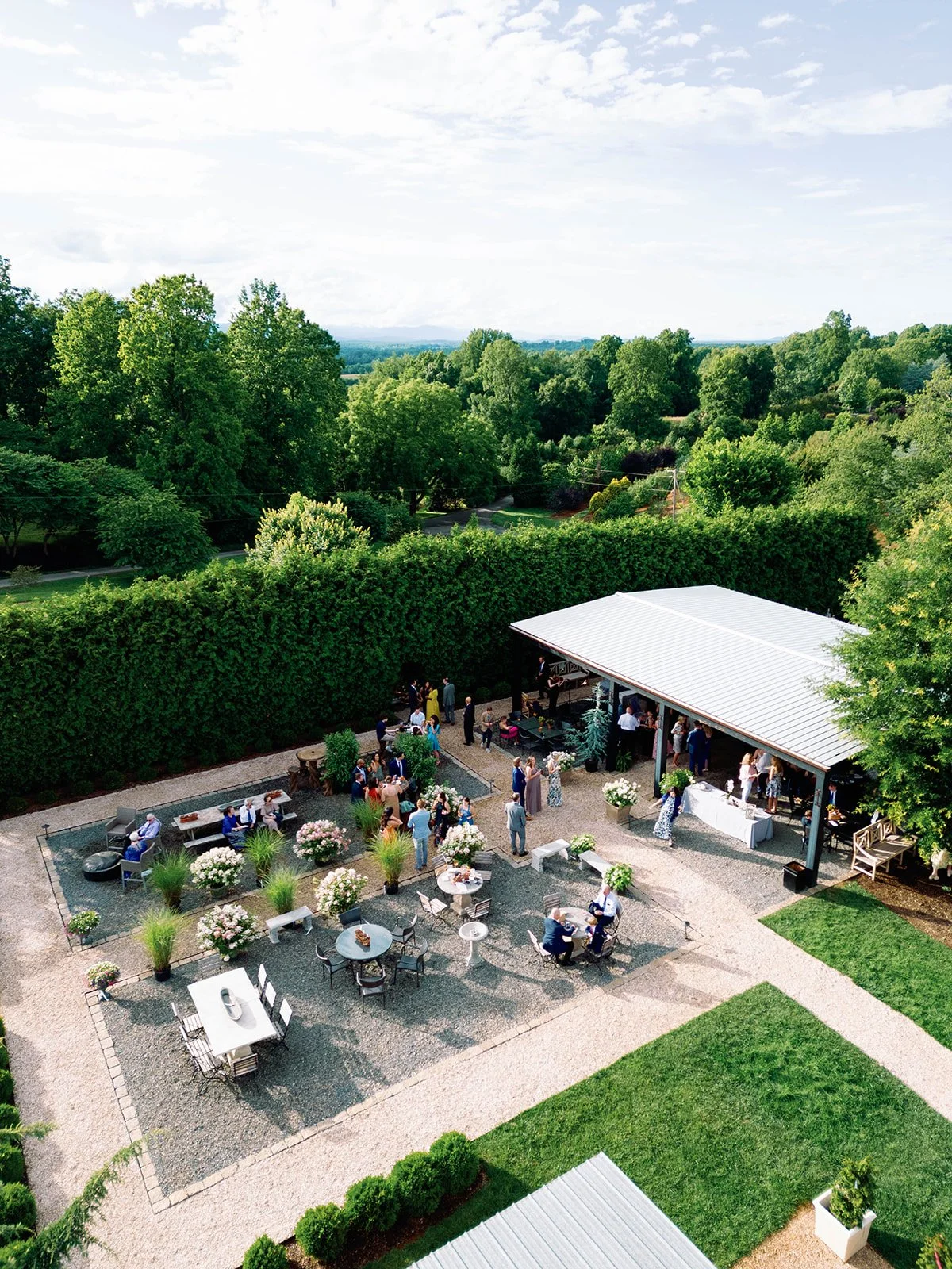 An outdoor party or event in a garden setting with people socializing, seated around tables, and standing near a covered area, surrounded by lush green trees and a cloudy sky.