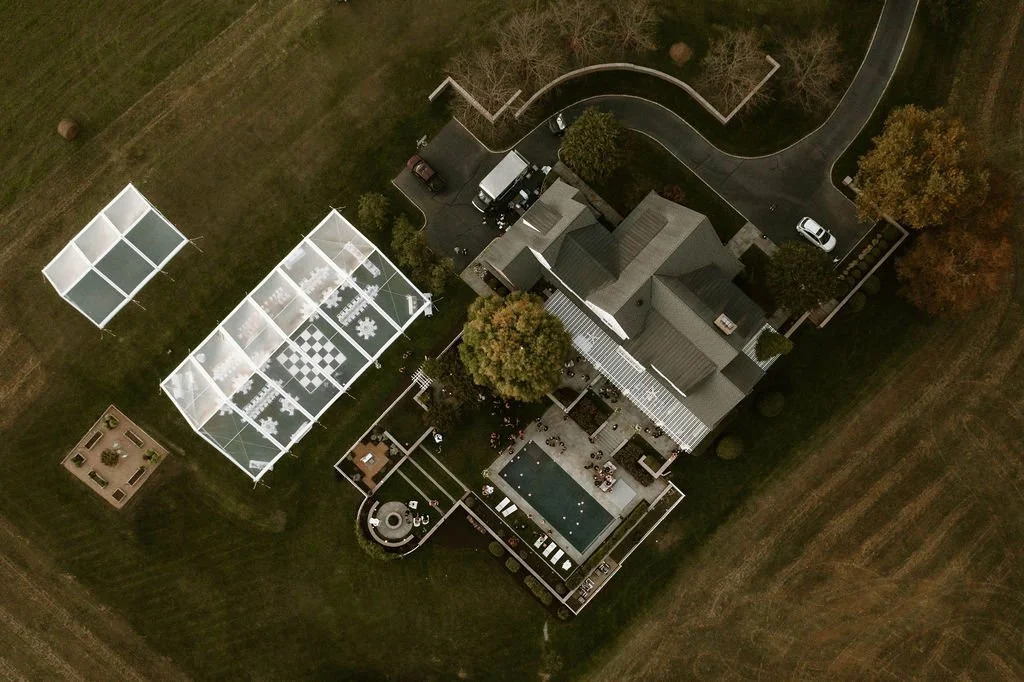 Aerial view of a large house with a backyard, featuring a pool, numerous outdoor seating areas, and a detached greenhouse with a glass roof, surrounded by a grassy lawn and trees.