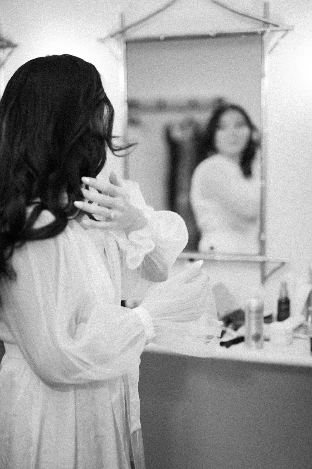 A woman with long dark hair looking at herself in a mirror while touching her neck, in a room with a dressing table and mirror.