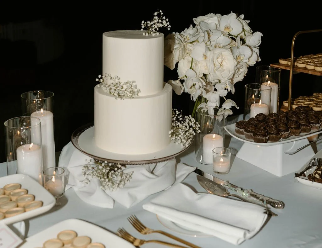 White two-tier wedding cake with small white flowers, surrounded by white flowers, candles, and assorted desserts on a decorated table.