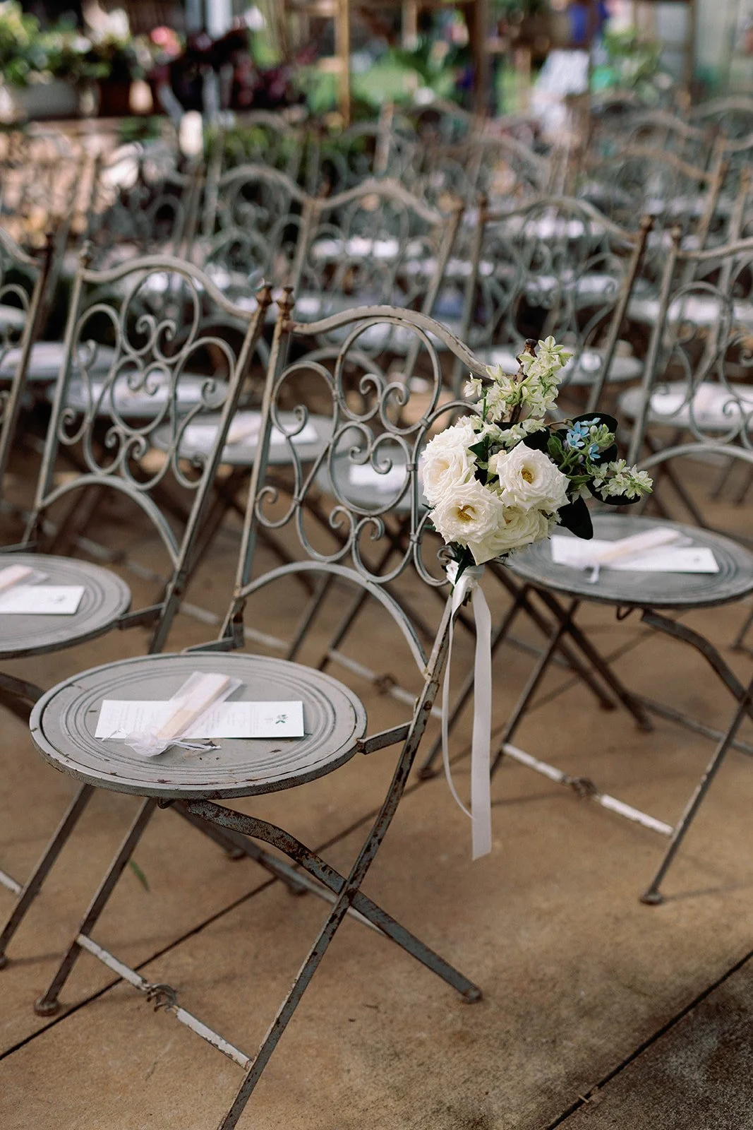 A row of metal chairs with scrollwork, decorated with a bouquet of white and light blue flowers, set up for a wedding or event.