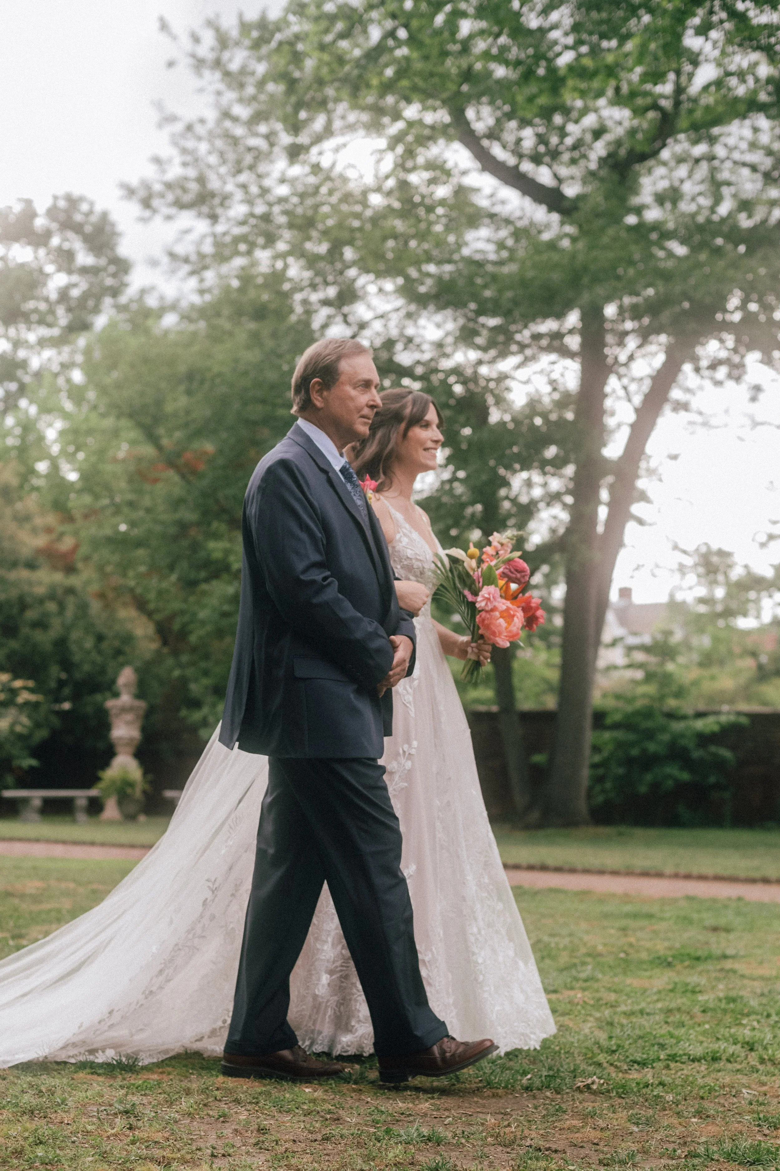 A bride in a white wedding dress holding a bouquet of pink, orange, and yellow flowers walking outdoors with an older man in a dark suit.