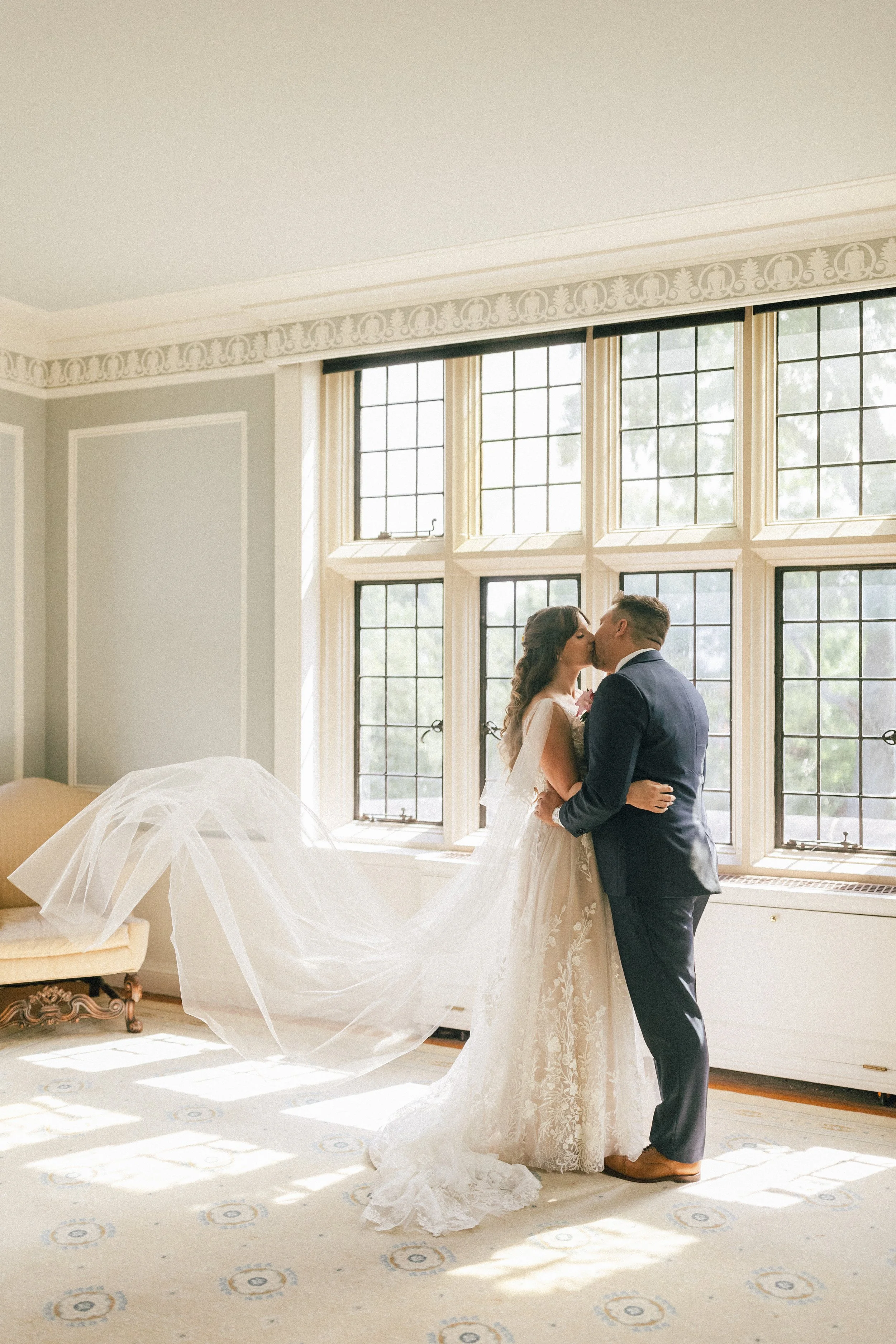 Bride and groom sharing a kiss indoors near a large window with sunlight streaming in, dressed in wedding attire.