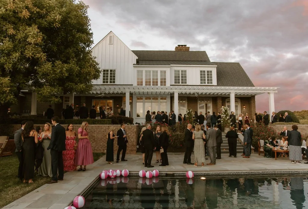 A large gathering of people dressed in formal attire at an outdoor event near a white house with a pool and pink and white balloons in the water, during sunset.