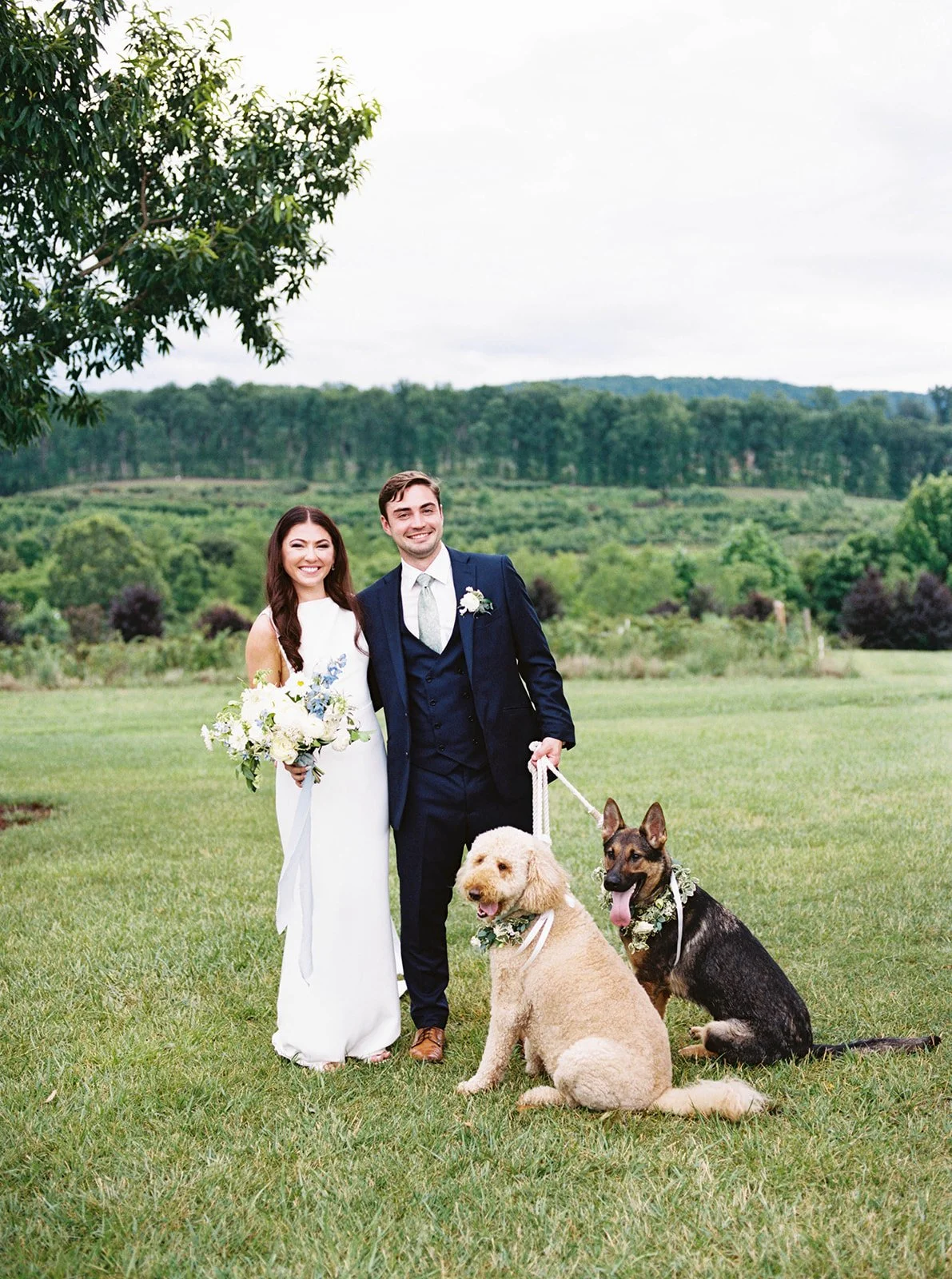 A bride and groom smiling outdoors on their wedding day, standing on a grassy field with two dogs that are wearing floral collars, with a scenic background of trees and rolling hills.