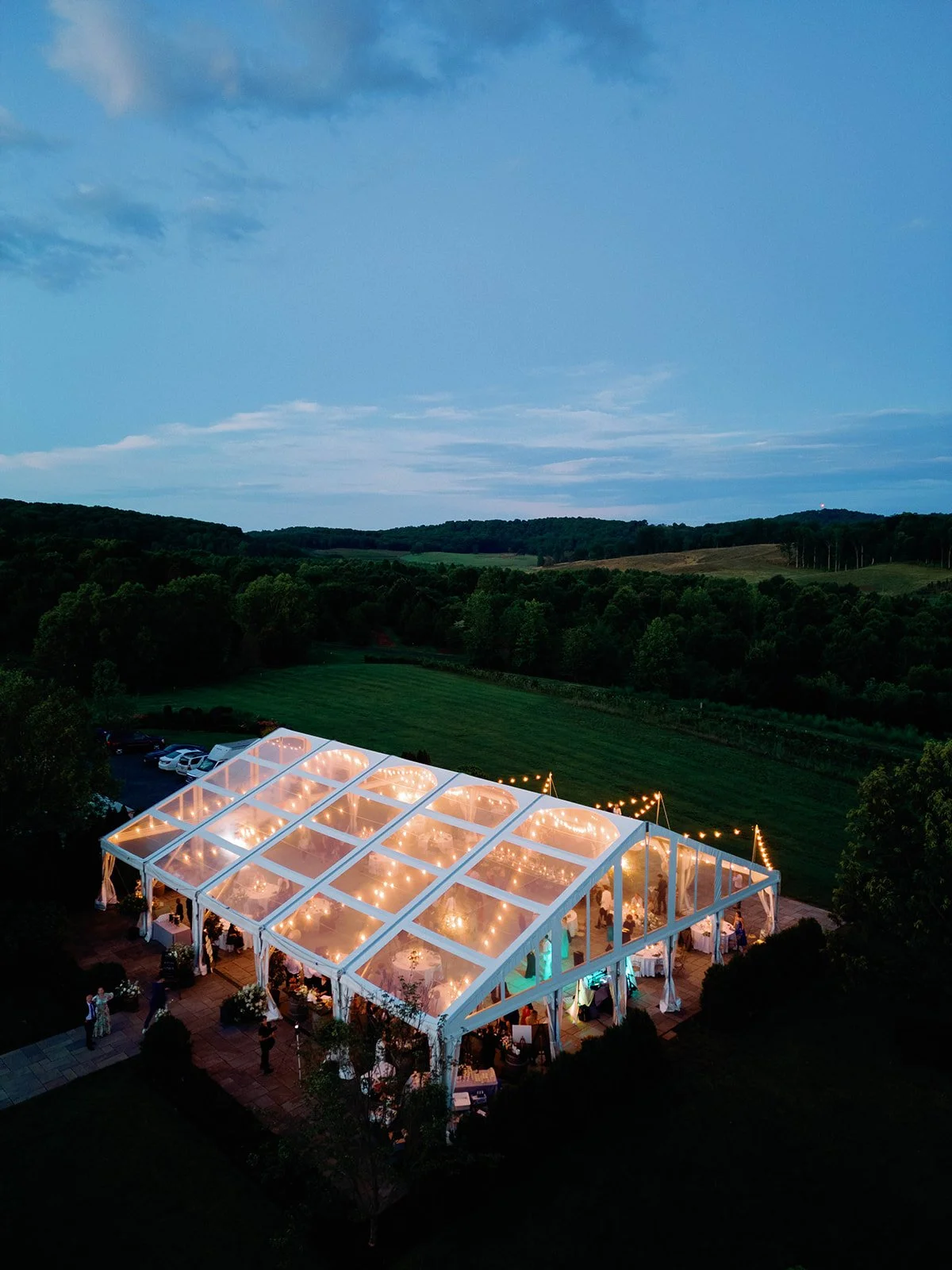 An outdoor event tent with string lights at dusk, surrounded by trees and open fields.