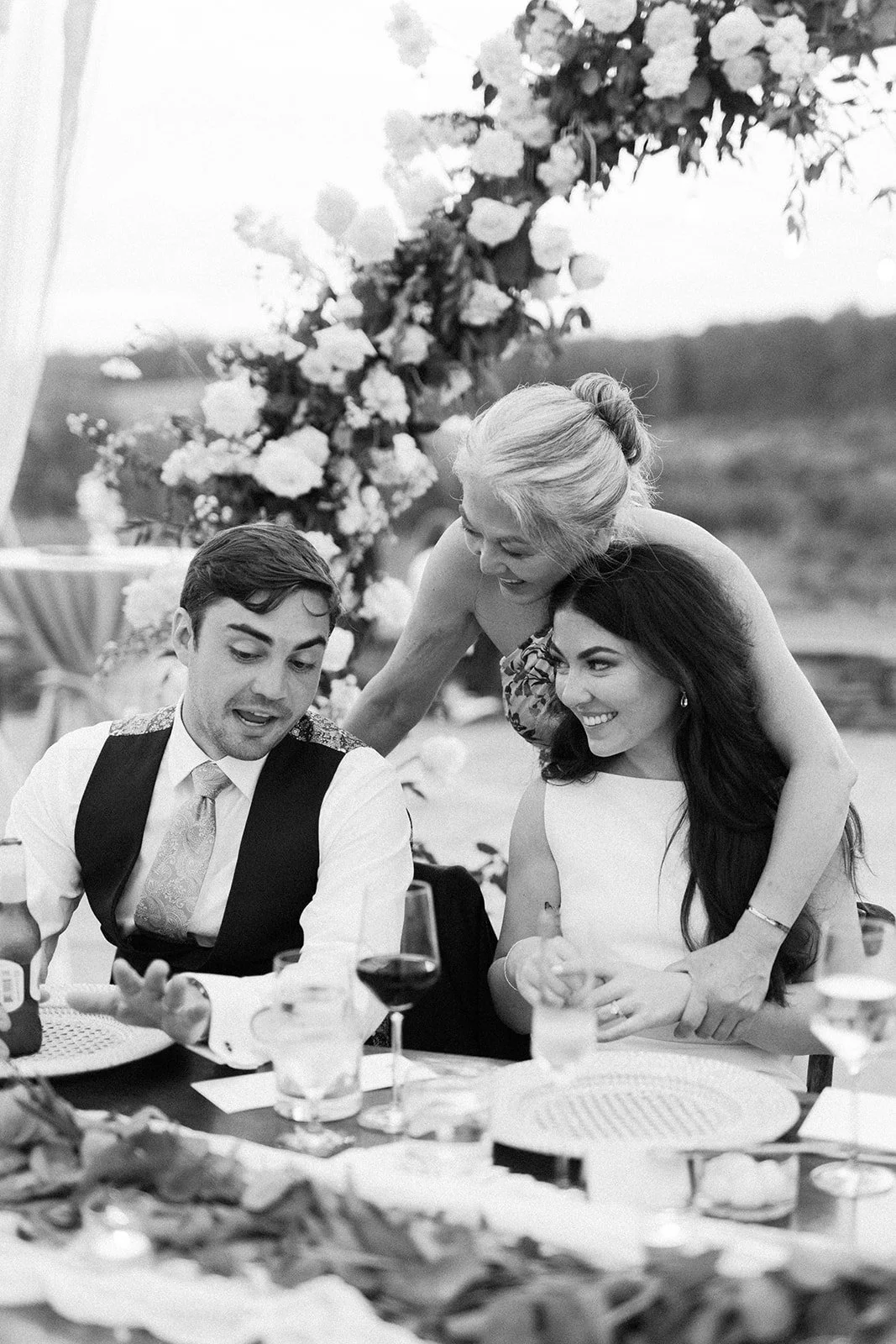 A black-and-white photo of three women at a wedding reception, smiling and engaging in conversation. One woman stands behind a seated couple, leaning over and smiling while the couple looks at something on the table. The scene includes a decorated ba