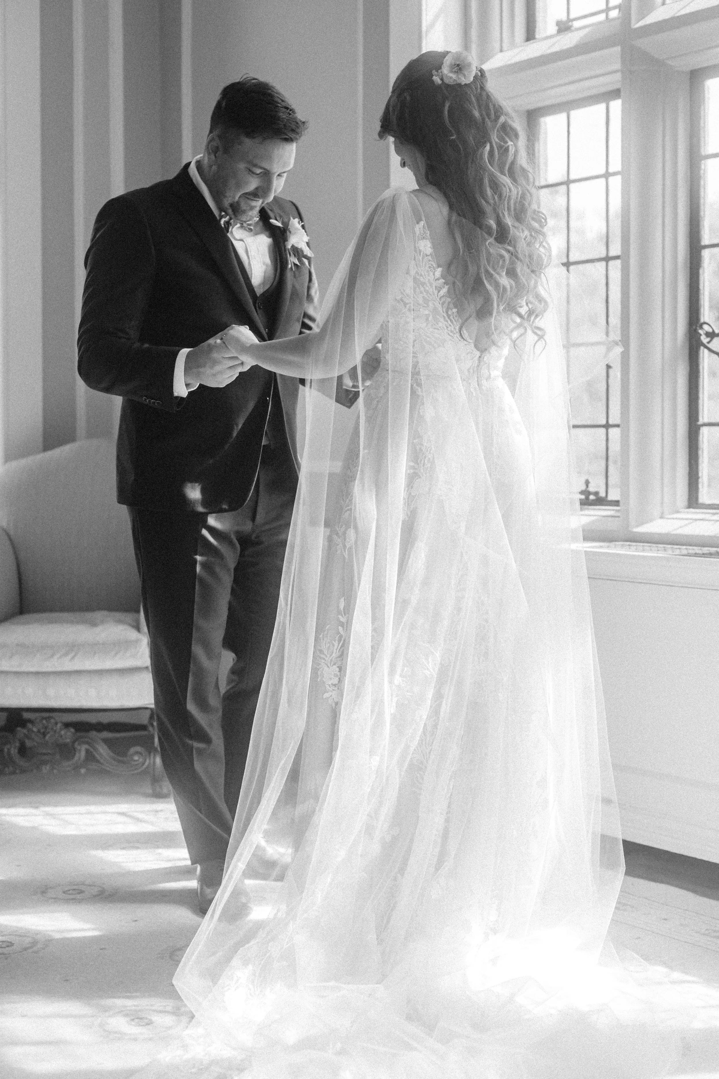 A black-and-white photo of a bride and groom exchanging vows indoors next to a large window, with the groom placing a ring on the bride’s finger.