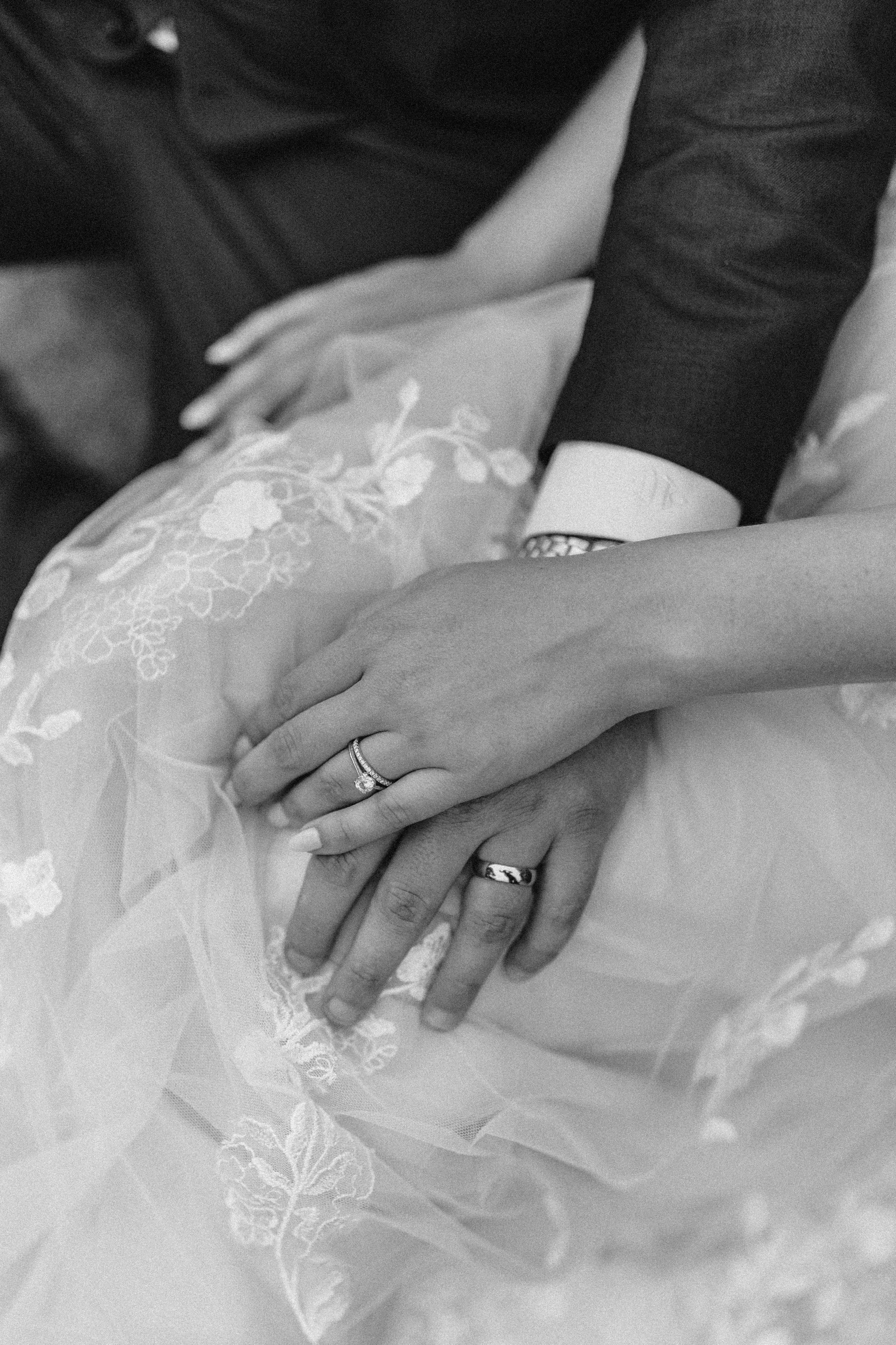 Close-up of a bride and groom's hands showing wedding rings, with the bride wearing an embroidered wedding dress and the groom wearing a suit.