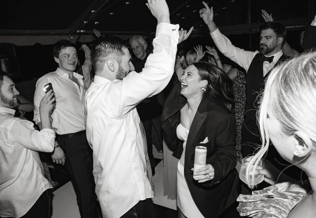 People dancing and celebrating at a party, smiling and raising hands, with some holding drinks.