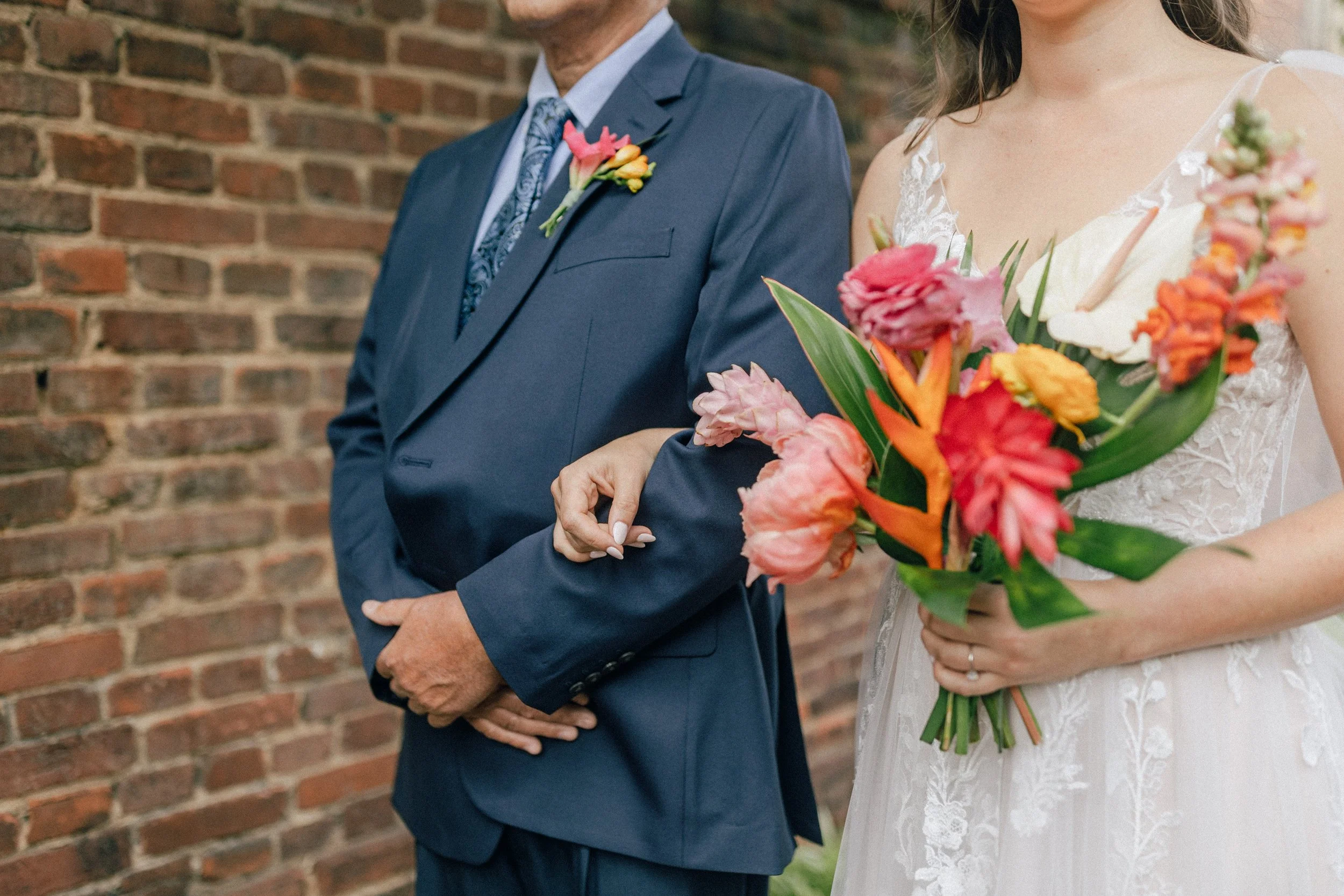 A bride dressed in white holding a bouquet of pink, orange, and green flowers, standing next to a man in a blue suit with a floral boutonnière, against a brick wall.
