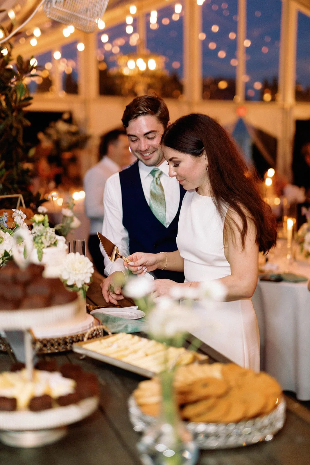 A man and woman at a wedding reception preparing to cut a wedding cake, surrounded by other guests and desserts, with string lights and candles creating a warm atmosphere.