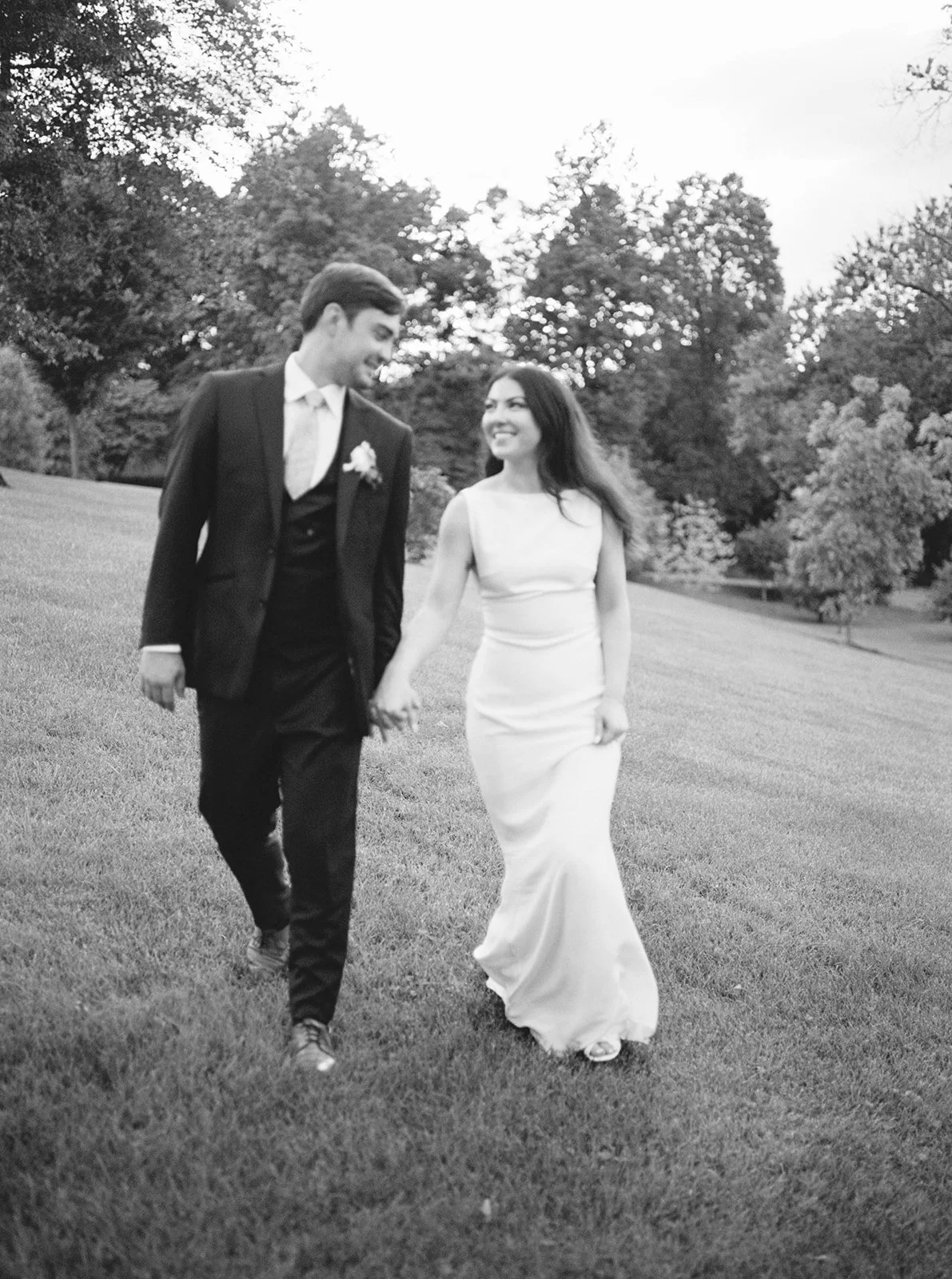A black-and-white photo of a bride and groom walking hand in hand outdoors, smiling, with trees and a grassy field in the background.