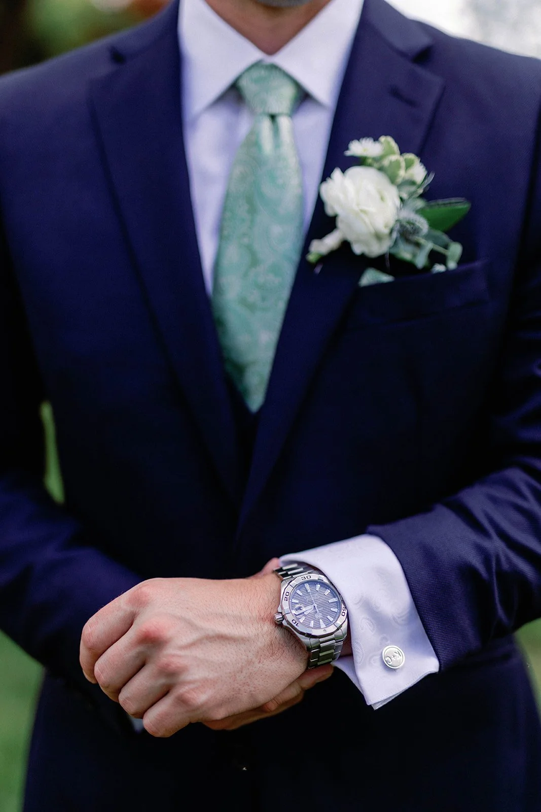 Close-up of a man in a navy suit with a white shirt, green paisley tie, and a boutonniere, showing his silver wristwatch.