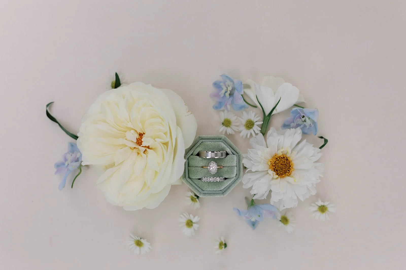 A set of three rings in a square green velvet ring box surrounded by white and light blue flowers and small white daisies on a light background.