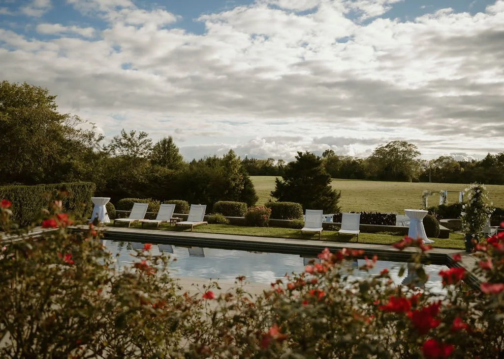 Swimming pool surrounded by white lounge chairs and garden flowers, with trees and a cloudy sky in the background.