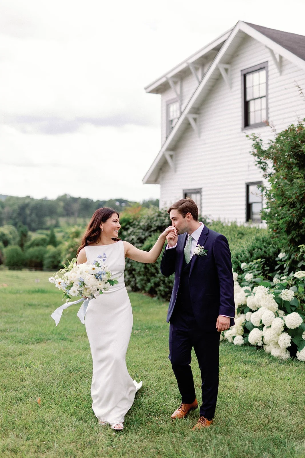 A bride and groom outdoors near a white house with green landscape in the background, the bride touching the groom's face as he kisses her hand, the bride holding a bouquet of white and purple flowers.