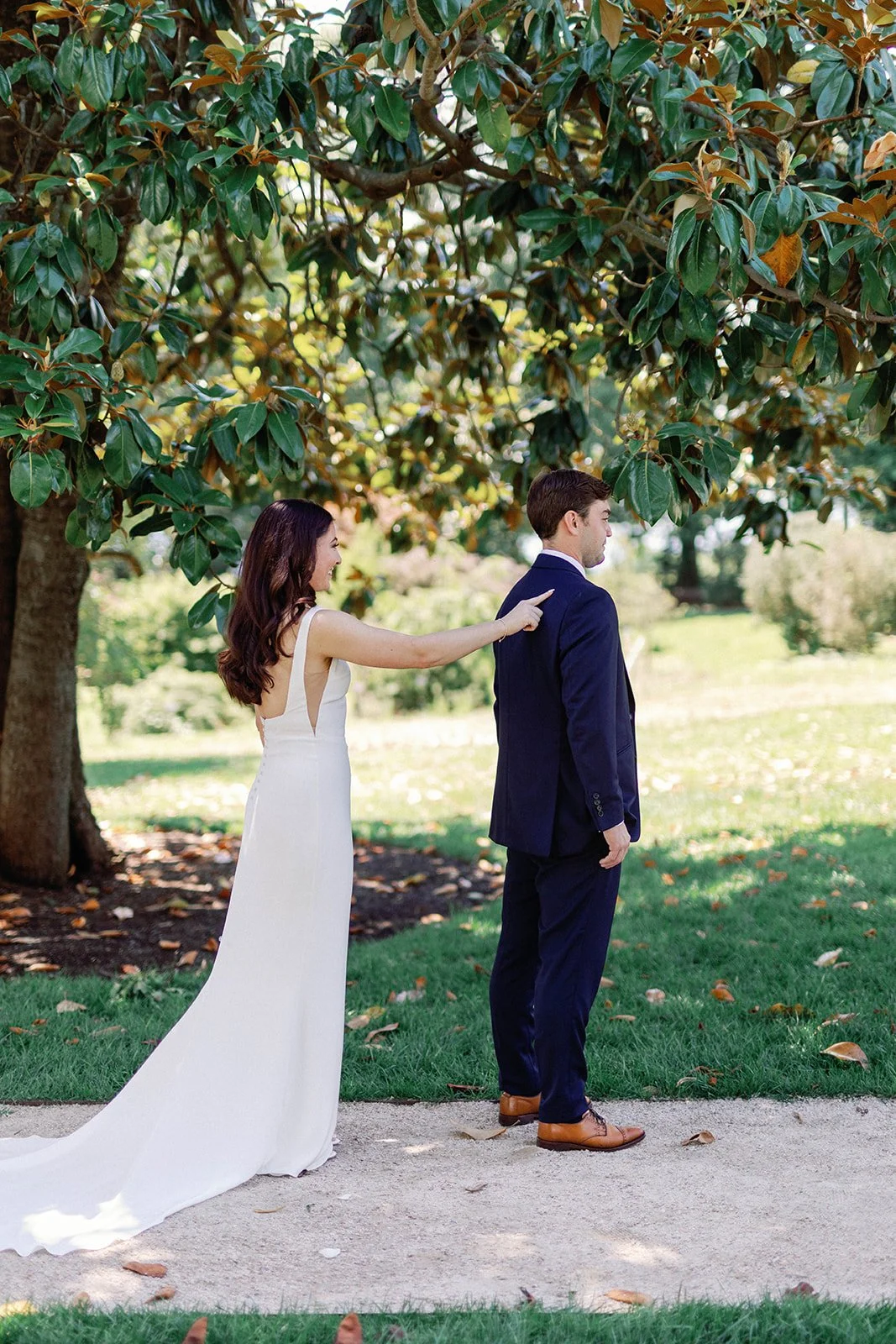 A woman in a white dress tugging on a man's suit jacket from behind, in a park setting with trees and greenery.