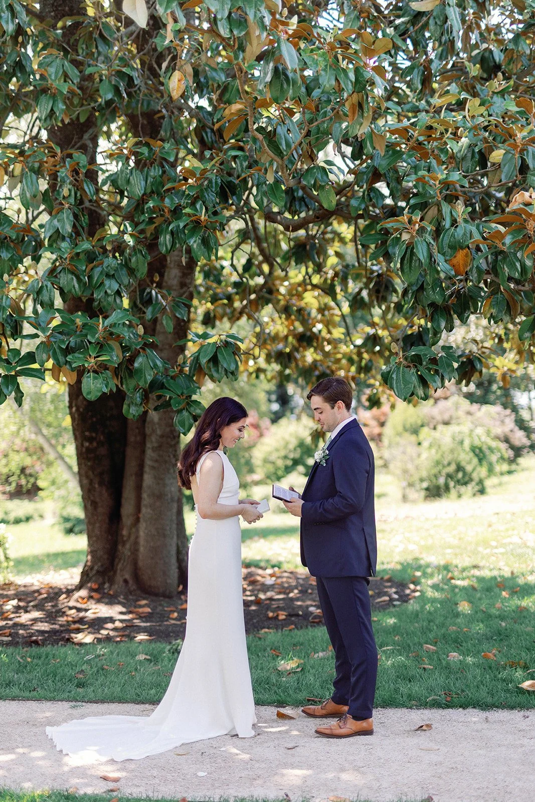 A bride and groom exchanging vows outdoors under a large leafy tree, with grass and fallen leaves on the ground in a sunny setting.