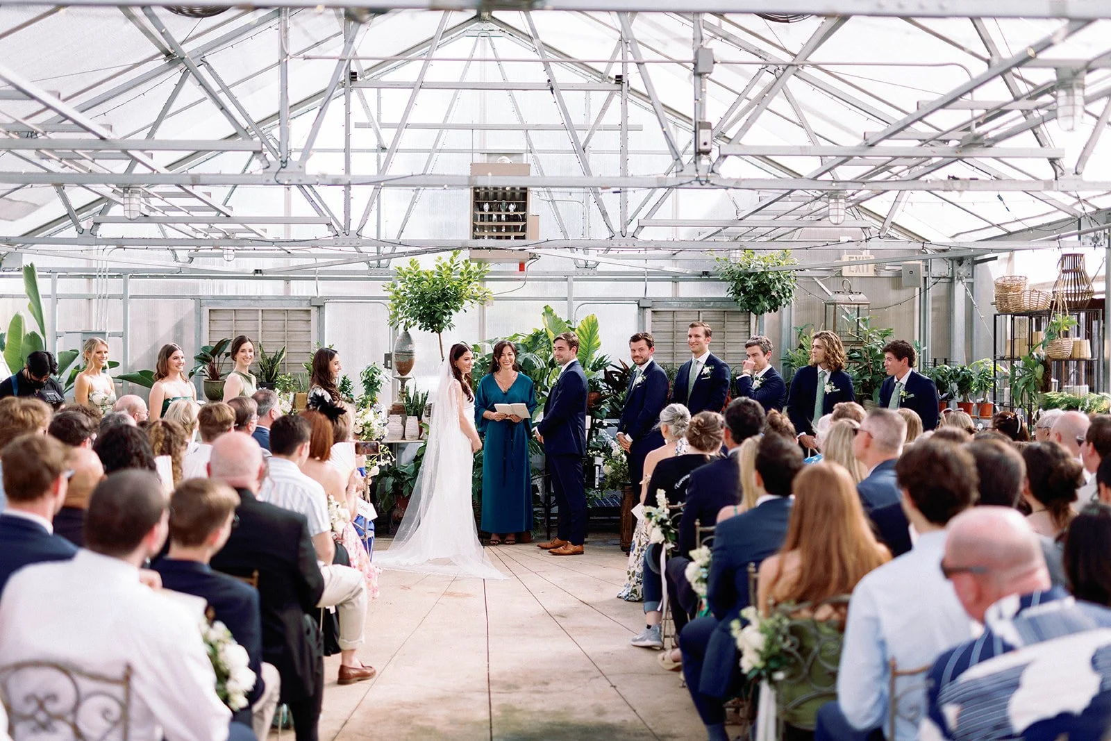 A wedding ceremony inside a greenhouse with guests seated and the bride and groom standing in front of officiant, surrounded by bridesmaids and groomsmen, and lush green plants.