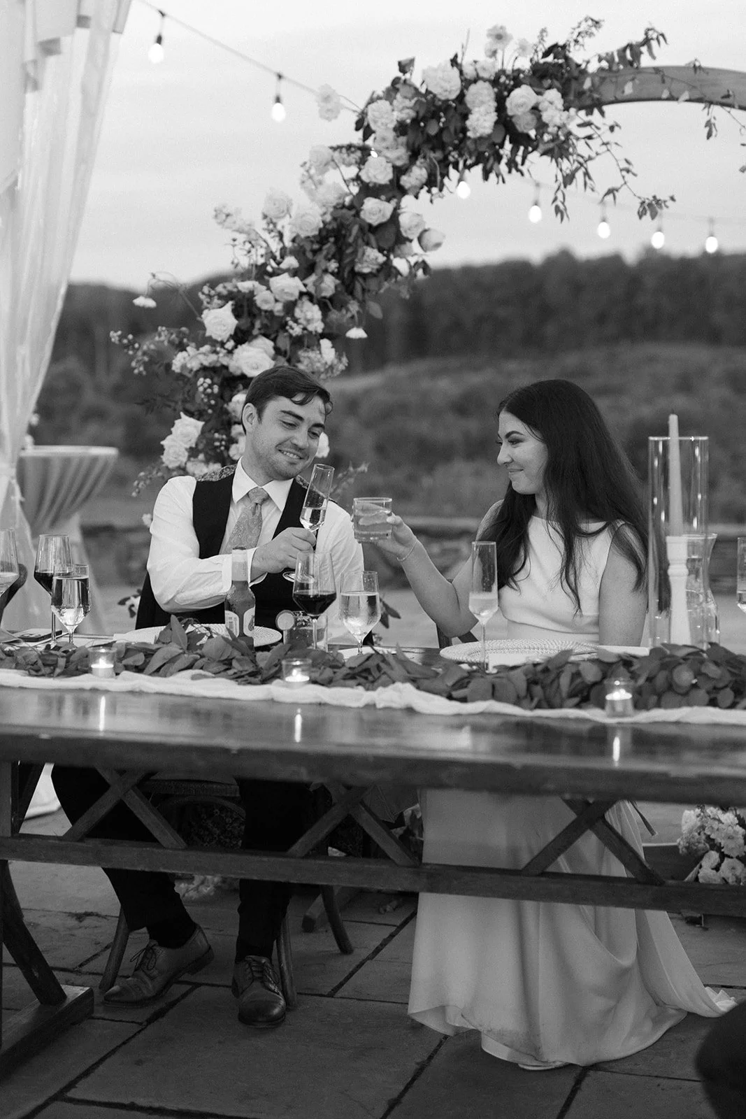 A couple at a wedding reception, smiling and toasting glasses, seated at a decorated table outdoors with flowers and candles, while arching floral decoration and string lights are visible in the background.