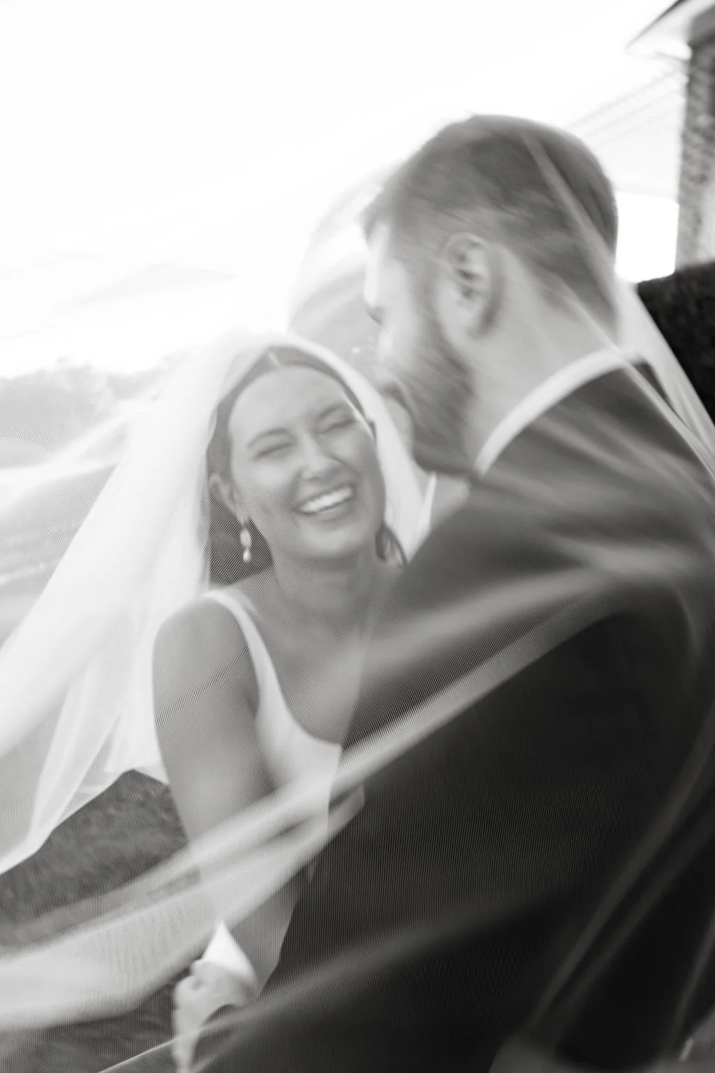 Black and white photo of a bride and groom embracing and smiling through a car window.
