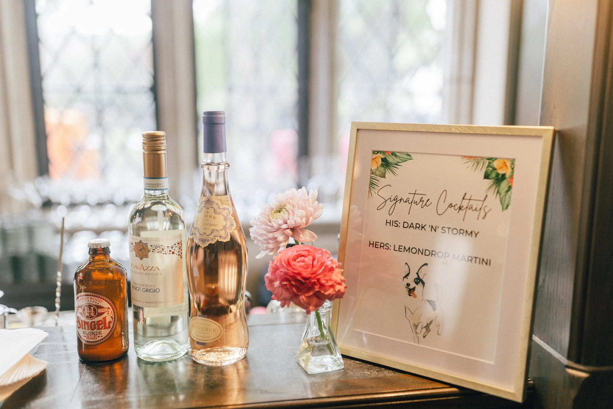 Table with bottles of alcohol, a small vase with pink and white flowers, and a framed sign listing signature cocktails, including a dark 'n' stormy and a lemonday martini.