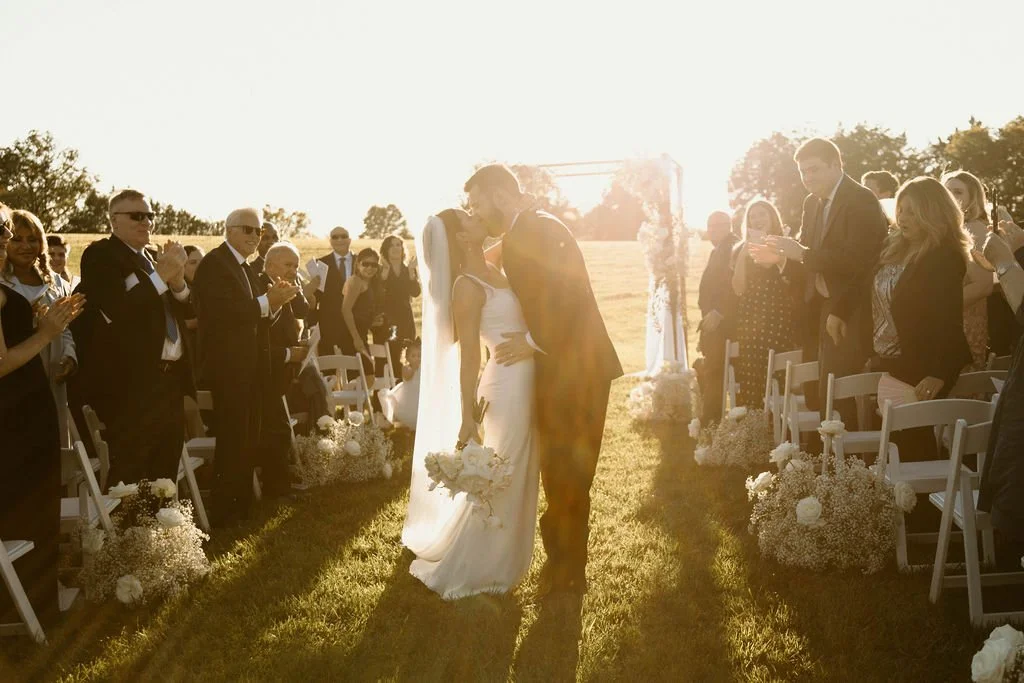 A bride and groom kissing at an outdoor wedding ceremony during sunset, with guests clapping and taking photos, decorated with white flowers.