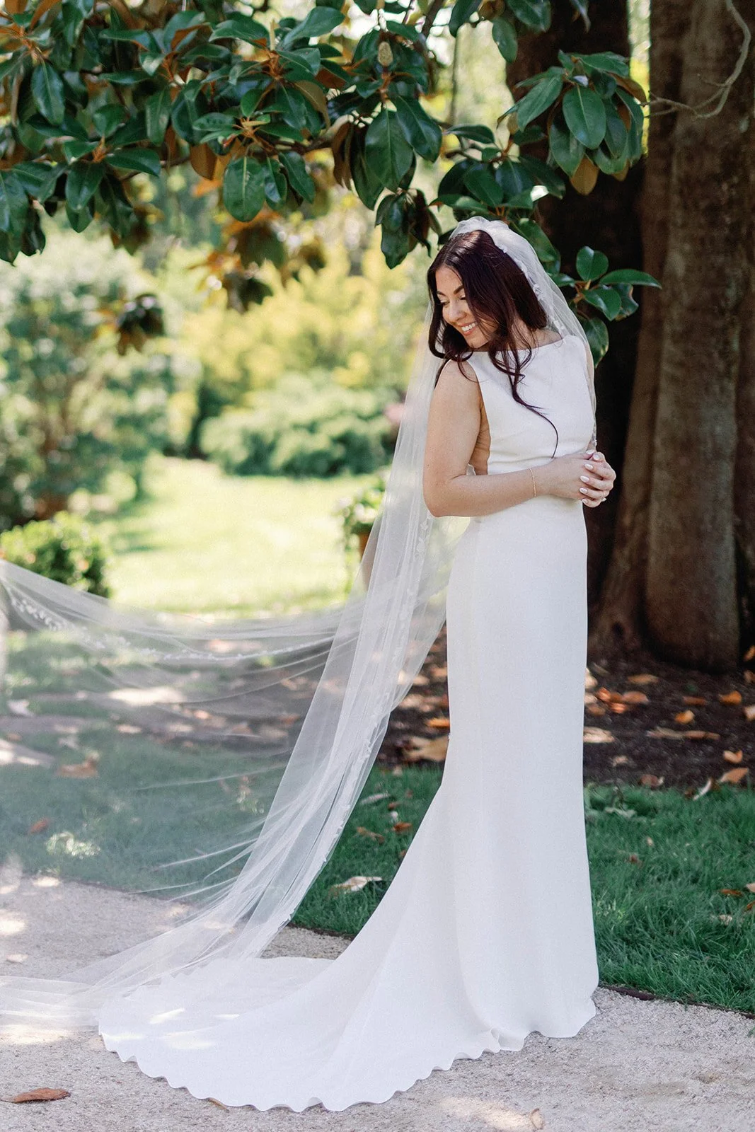 A bride in a white wedding dress with a long veil standing outdoors under a large tree, smiling and looking down, with greenery in the background.