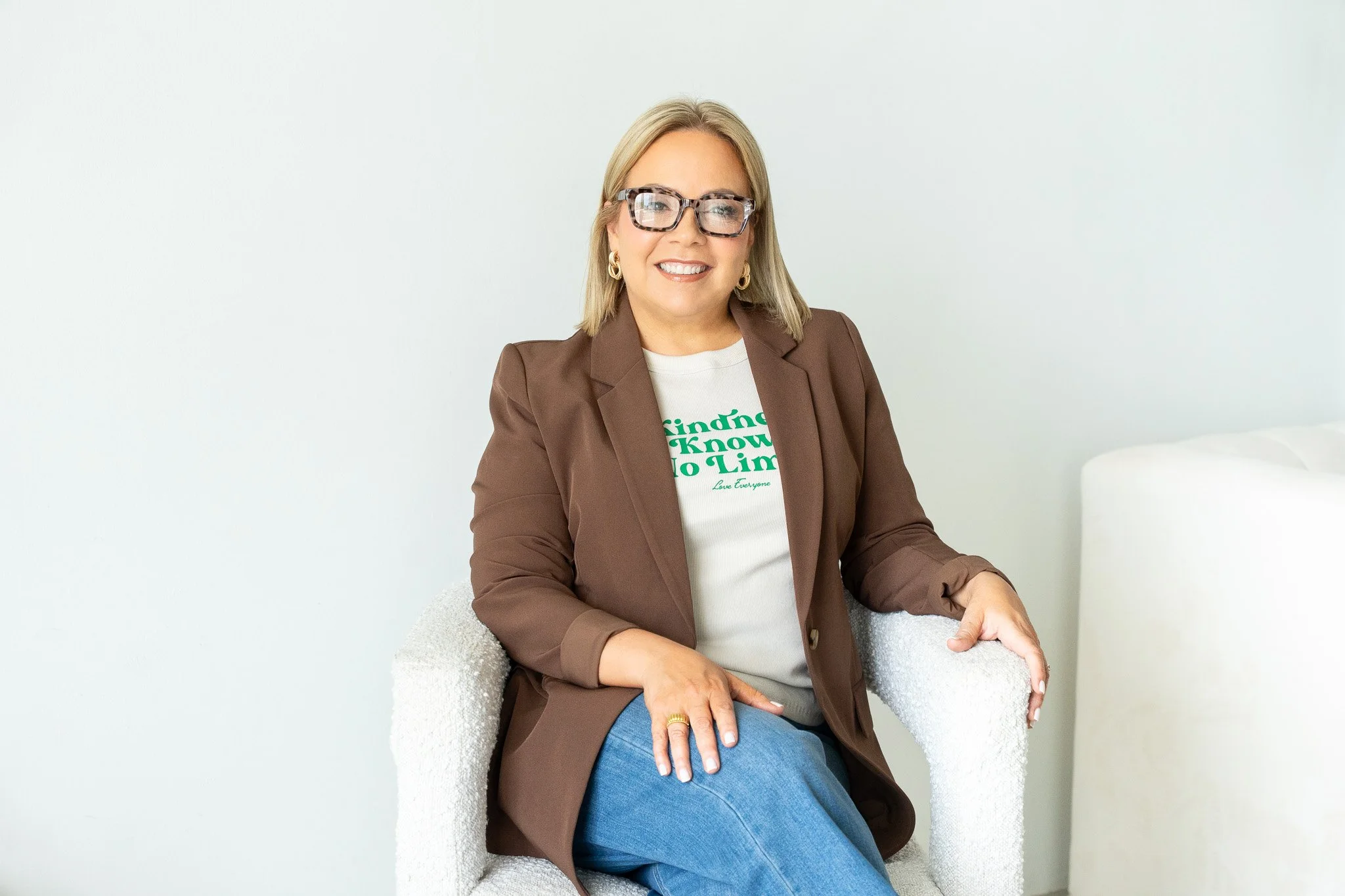 A woman with blonde hair, glasses, and earrings sitting in a white armchair, smiling at the camera, wearing a brown blazer and a white T-shirt with green text.
